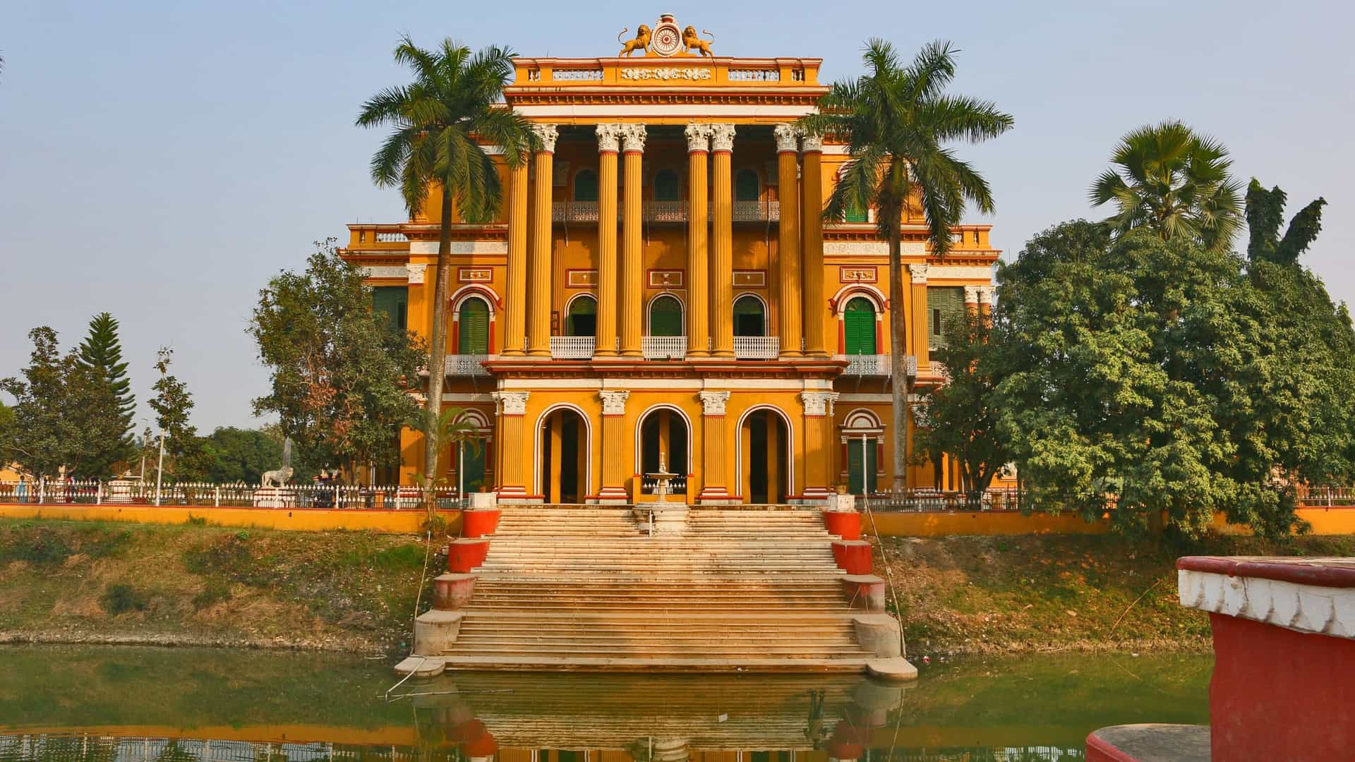 A stunning full-frontal view of the grand Murshidabad Palace, an impressive yellow historical building with tall pillars and balconies, situated on the bank of a river or large pond.