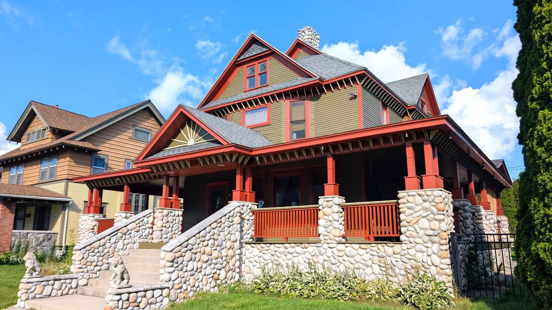 A historic two-story home in Muskegon, Michigan, has a prominent front porch, red trim, and a unique river rock foundation and staircase.