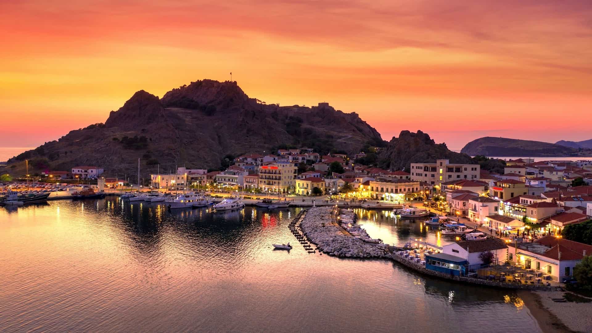 A vibrant sunset view of the harbor and town of Myrina, Greece, with boats docked and lights illuminating the buildings against a backdrop of a mountain with a castle ruin.