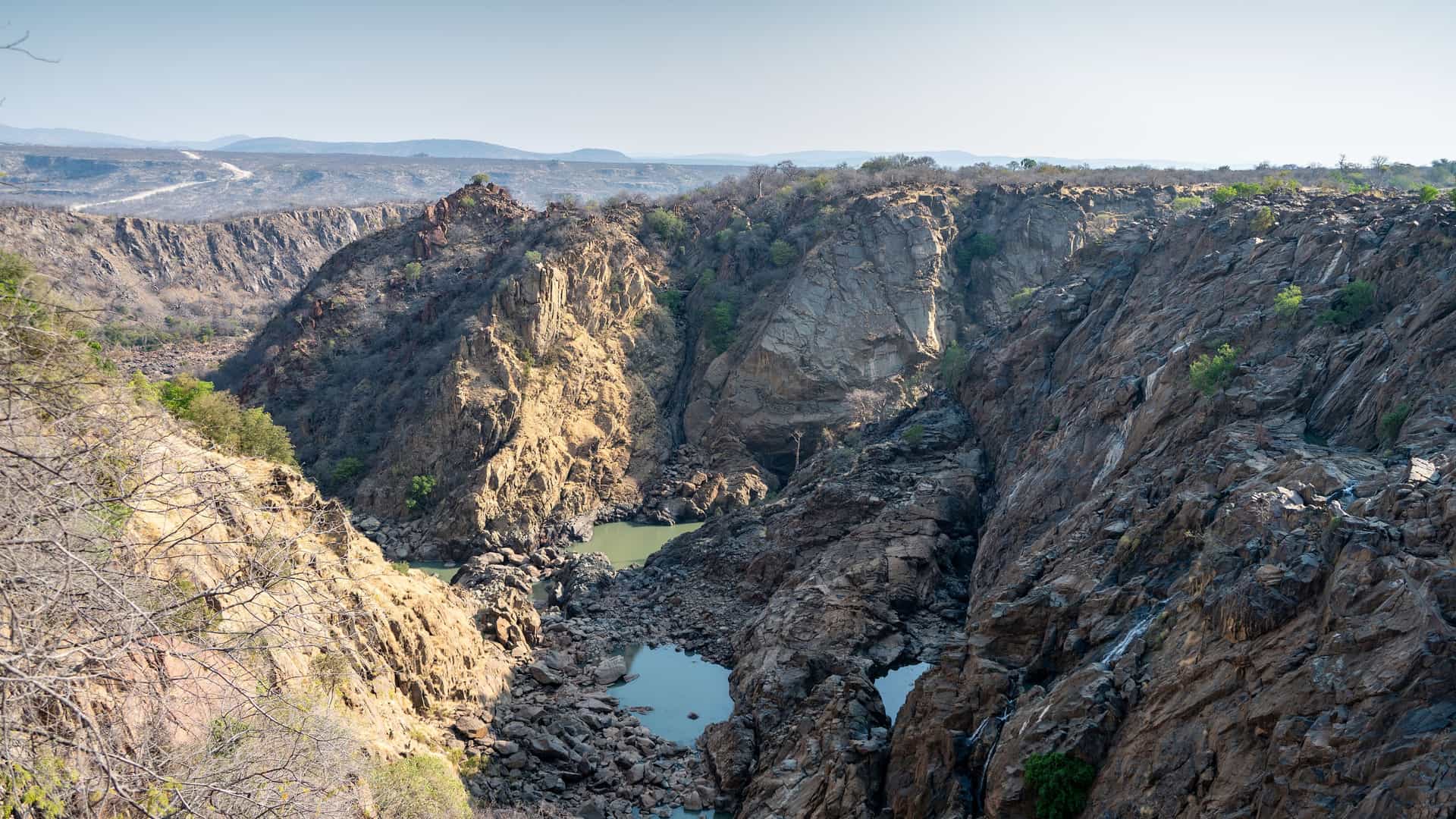 A stunning wide-angle view of the dramatic canyon landscape in Namibe, Angola, with towering rock formations and small pools of water nestled at the bottom. 