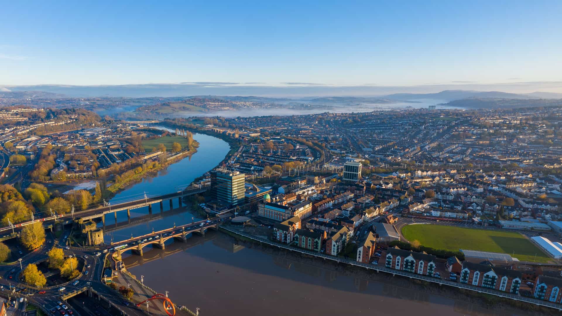 "An aerial panoramic shot of the city of Newport, Wales, with the River Usk winding through the urban landscape and a bridge connecting both sides of the city.  "