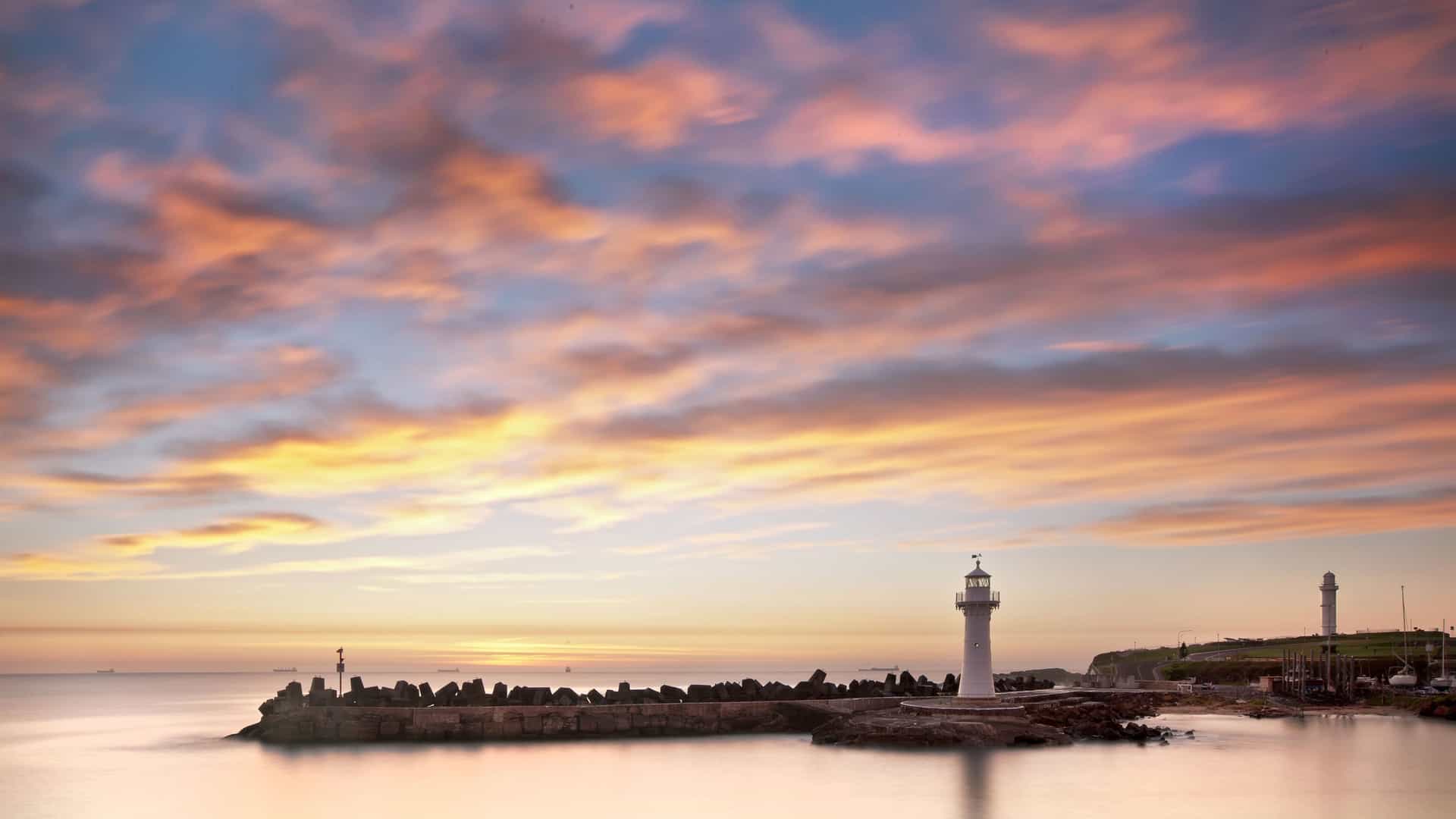 A stunning sunrise over a tranquil sea, with a lone lighthouse standing on a rock breakwater, with orange and yellow clouds on Norfolk Island.