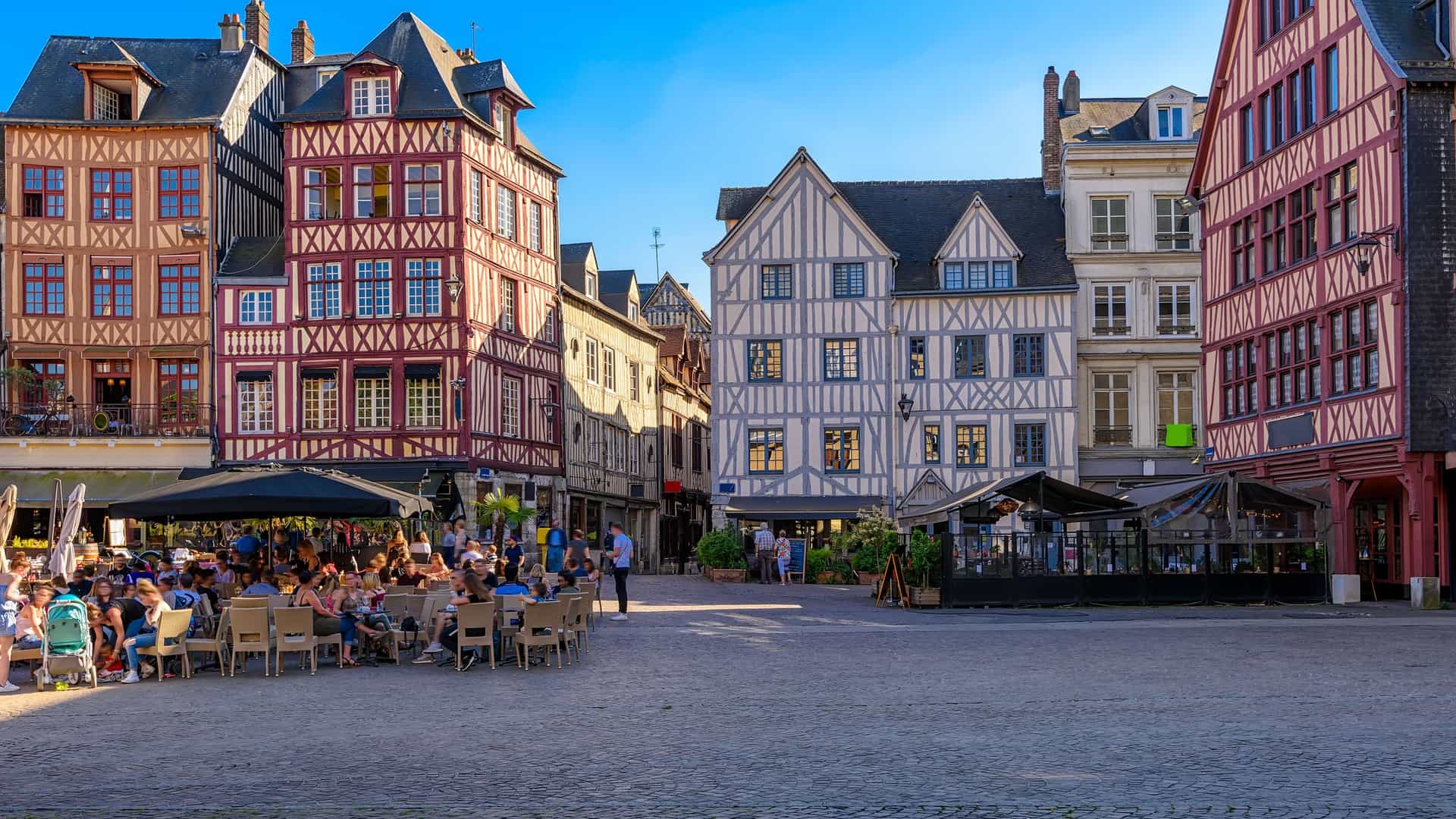 "A bustling summer day in Rouen, Normandy, with people dining at an outdoor cafe in front of historic, half-timbered houses in the Place du Vieux Marché.  "