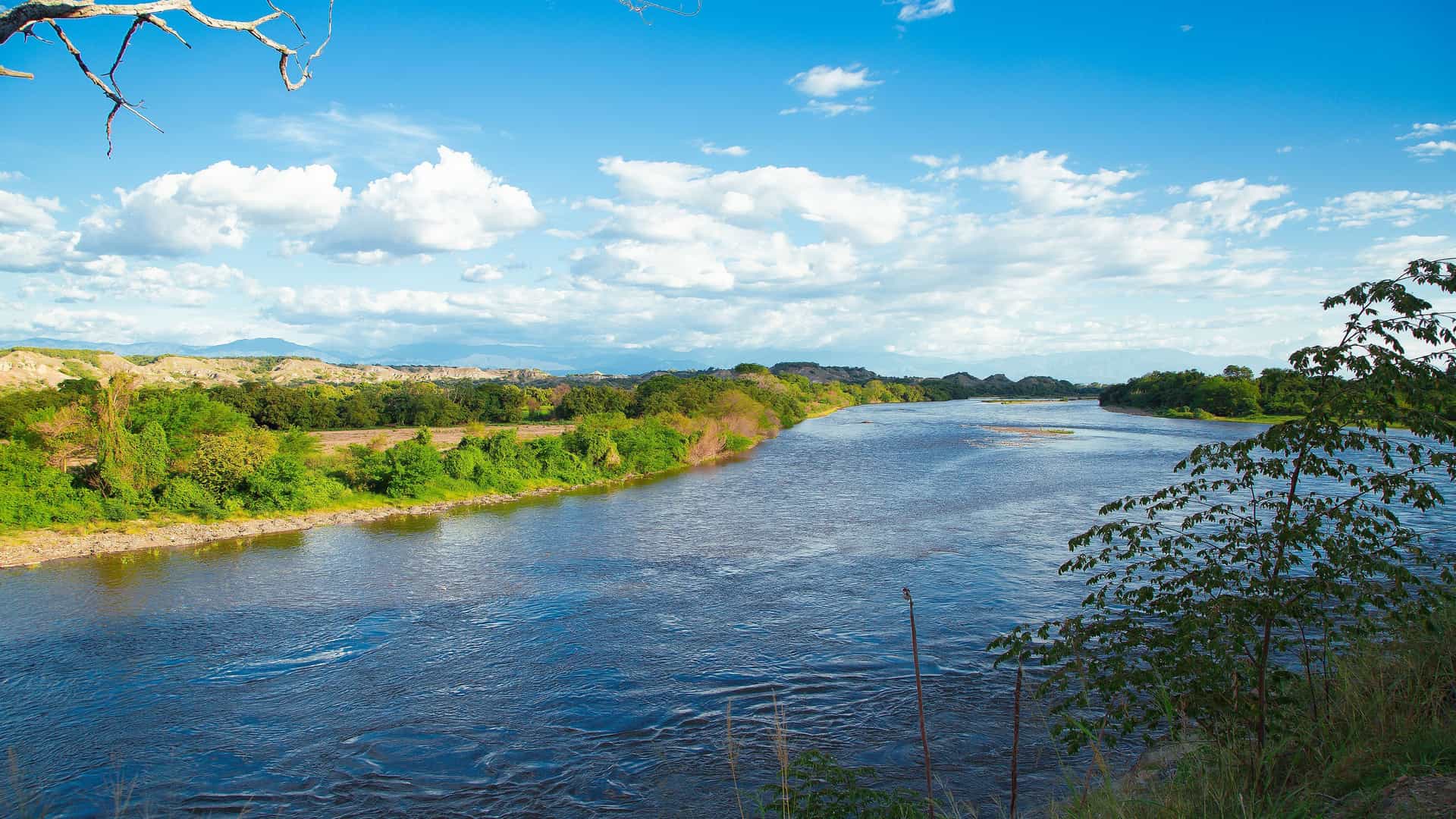 A panoramic shot of a wide, calm river flowing through a lush, green landscape under a bright blue sky with white clouds in Nueva Venecia, Colombia.