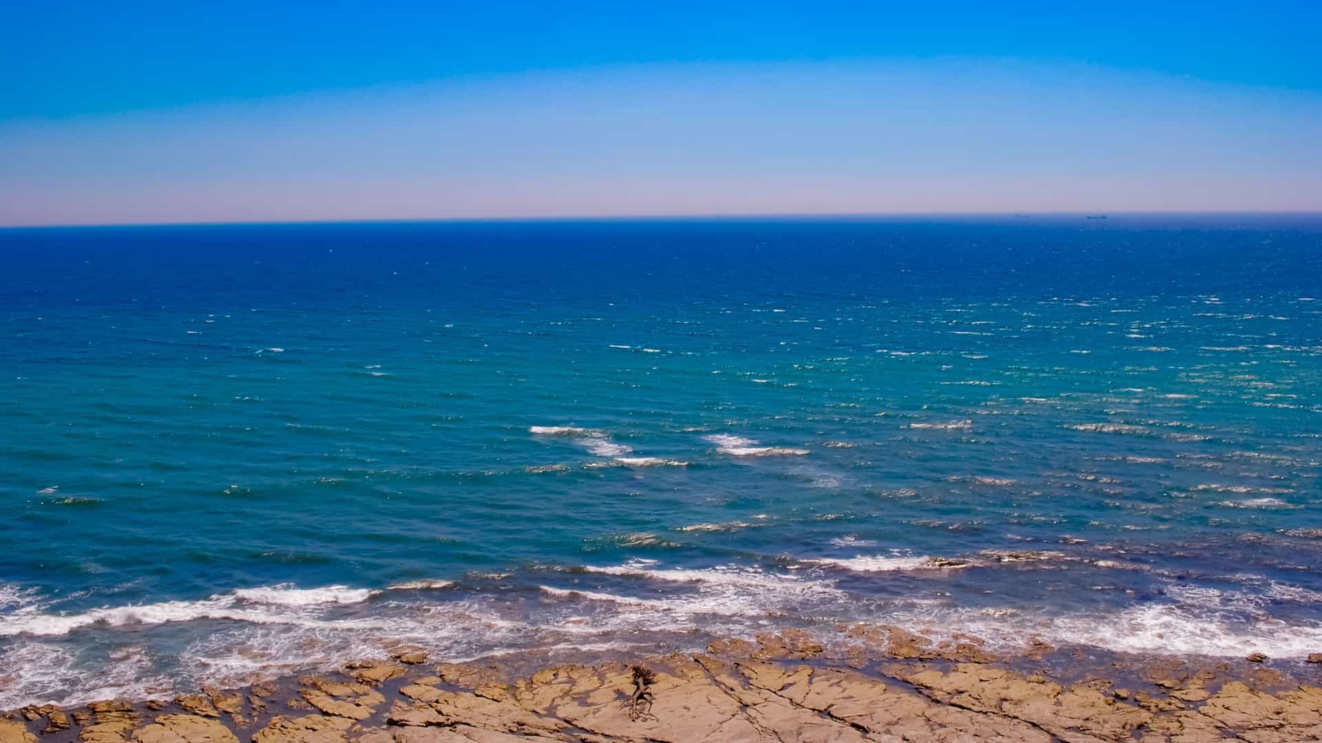 A scenic shot of the rocky coastline of Omaezaki, Japan, with waves crashing against the shore and the expansive blue Pacific Ocean extending to the horizon under a clear sky.