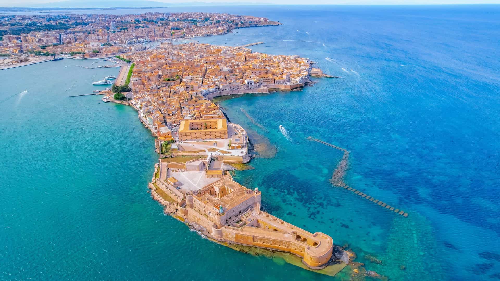The historic island of Ortigia in Syracuse, featuring the Maniace Castle and a densely built ancient city surrounded by turquoise water.