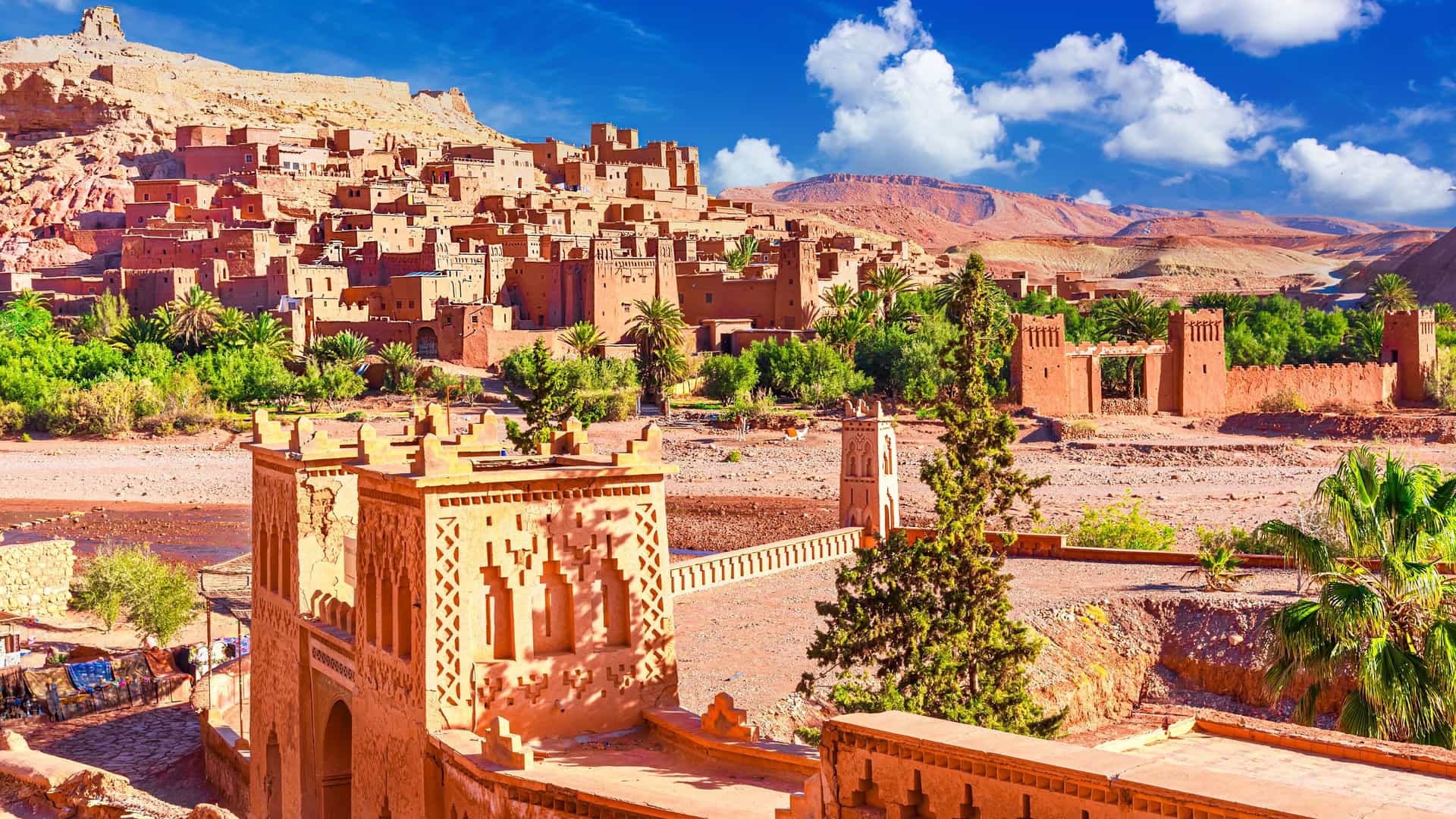 "A panoramic shot of the historic Aït Benhaddou, a fortified earthen city, or ksar, in Ouarzazate, Morocco, with palm trees and a lush green oasis at its base.  "