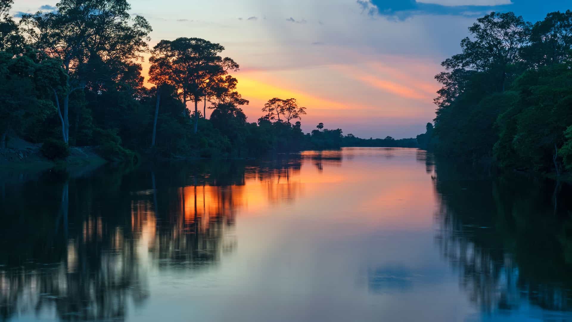 A wide shot of the tranquil Pacaya River in Peru, surrounded by a thick, lush jungle with the sunset reflecting on the calm water.