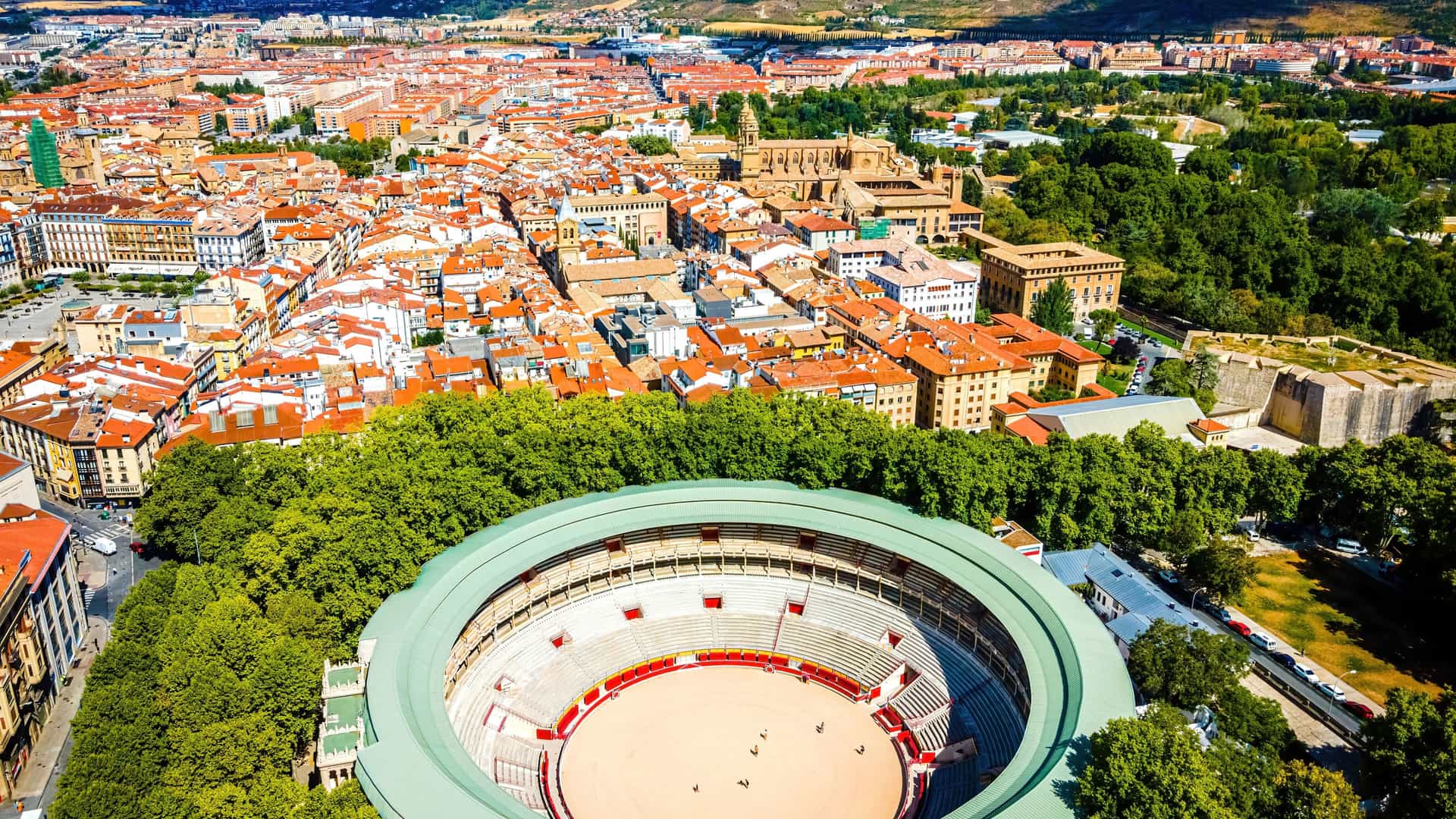 "An aerial panoramic shot of the historic Plaza de Toros, the bullring in Pamplona, Spain, with the city's old town and surrounding green trees and landscape in the background.  "