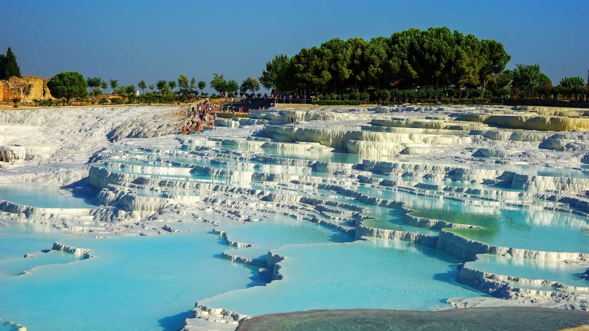 "A stunning wide-angle shot of the iconic Pamukkale thermal pools in Turkey, with people relaxing in the blue terraced waters and lush green trees in the background.  "