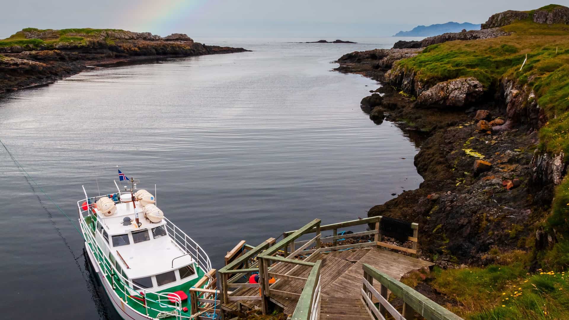 A scenic view of Papey Island, Iceland, with a boat docked in the rocky harbor and a rainbow faintly visible in the distance.