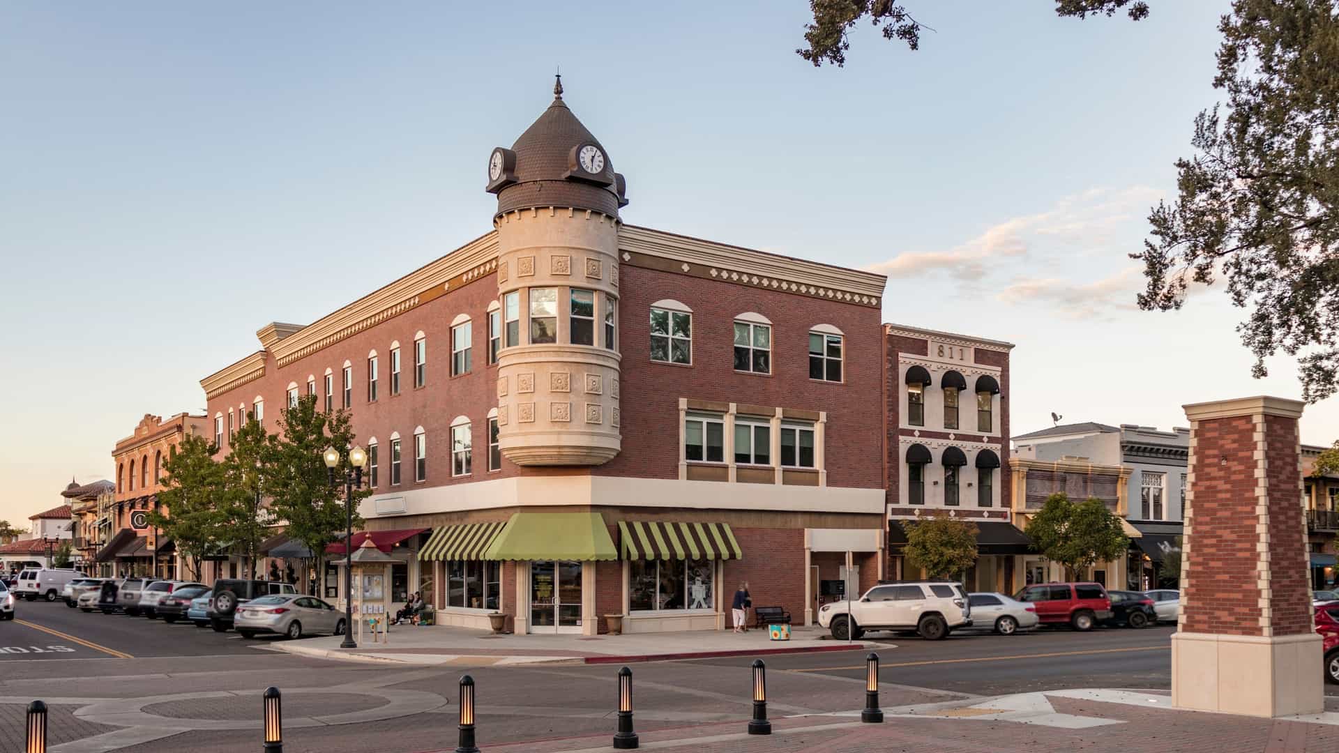 A horizontal shot of Downtown Paso Robles, Central California, USA taken at sunset. The photo is an eye-level view that captures the architecture along the main street, which is likely 12th Street or a street facing the city park. The streetlights and building lights are illuminated, and the windows reflect the warm, golden-hour light of the setting sun. Parked cars line the street, and there is visible pedestrian activity, giving the scene a vibrant, evening atmosphere.