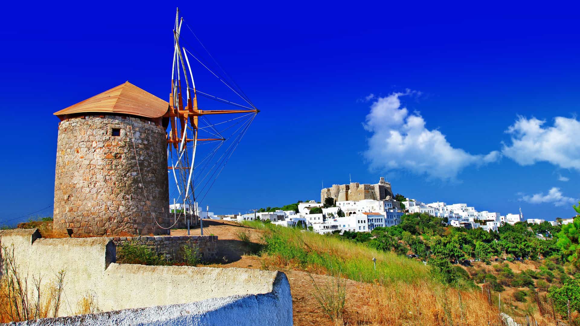 A traditional windmill with a pointed roof and wooden blades on a hill overlooking the white buildings of Patmos, Greece, with the historic Monastery of Saint John in the distance.