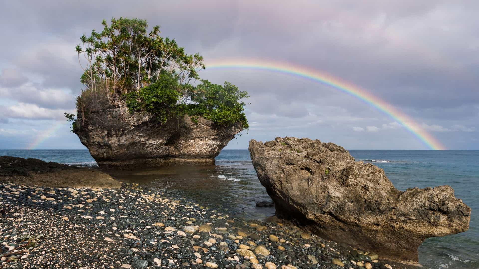 A view of a huge rock island covered in trees and another large rock in the foreground on the shore of Pentecost Island, Vanuatu, with a rainbow arching across the sky.