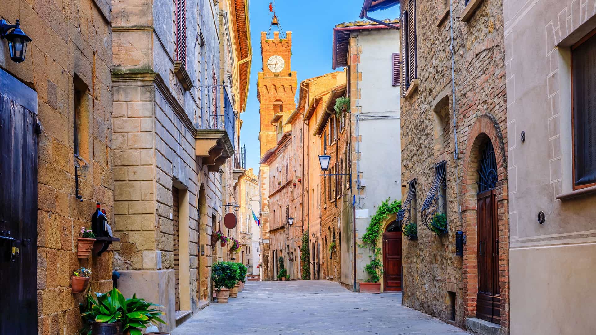 A view down a narrow, empty street in the town of Pienza, Italy, with ancient stone and brick buildings lining both sides. At the end of the street, a tall, medieval clock tower stands out against a bright blue sky.