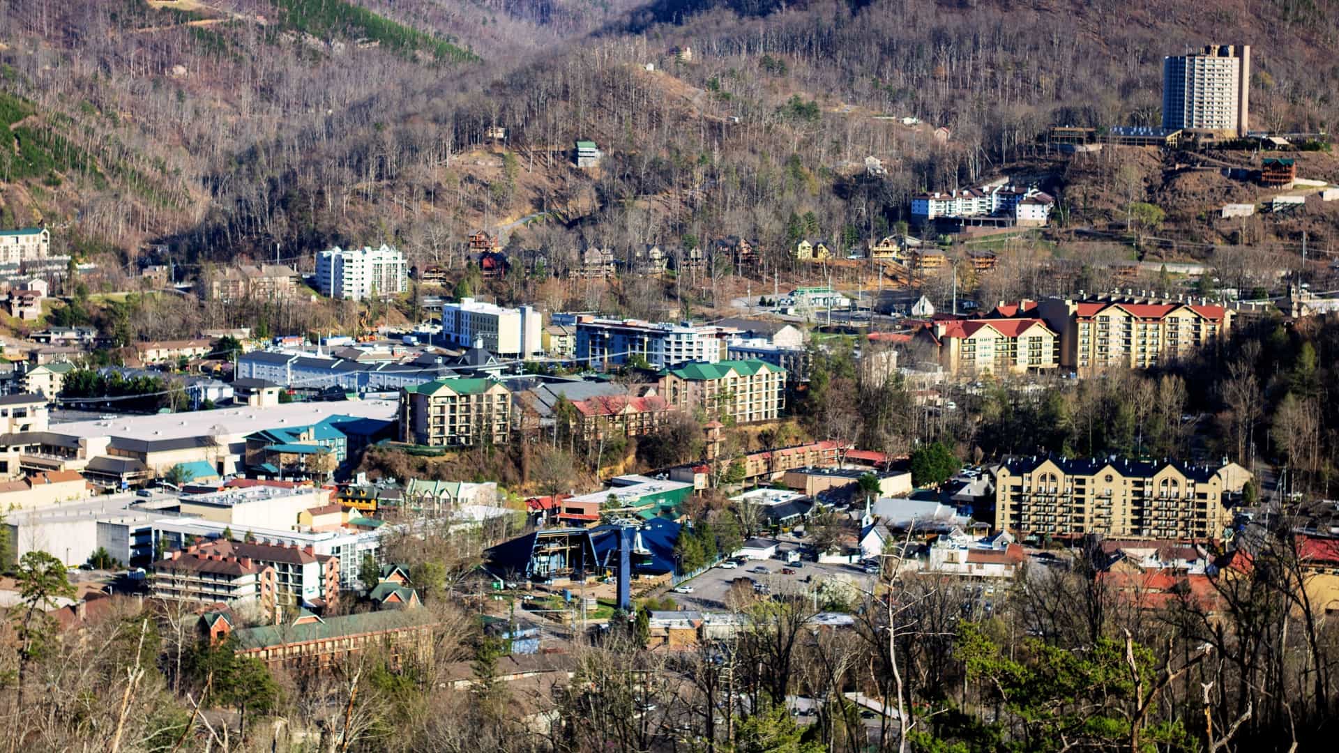 A high-angle or aerial overlook view of the city of Pigeon Forge, Tennessee. The photograph captures the bustling, low-rise commercial strip of the city's parkway, filled with various businesses and attractions, nestled in the valley floor against the dramatic, forested backdrop of the Great Smoky Mountains.