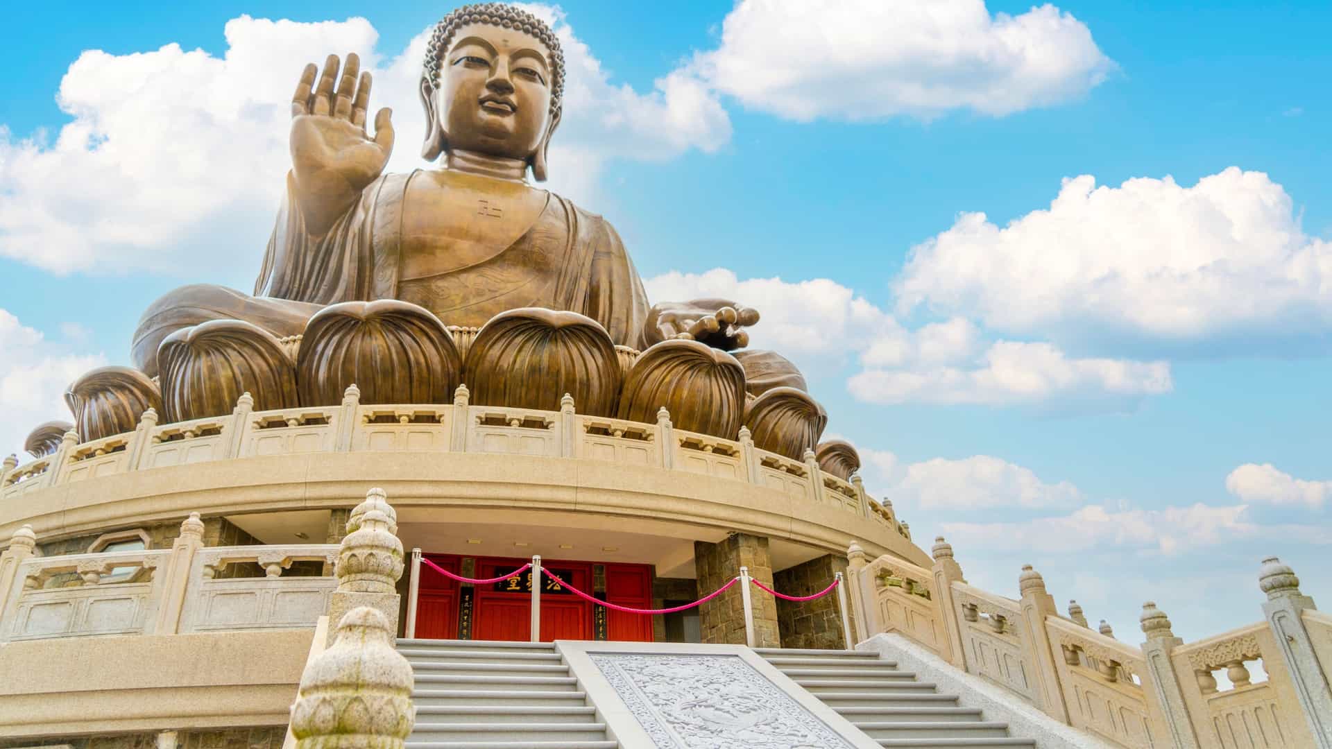 A towering bronze Tian Tan Buddha statue, sitting on a lotus throne, with one hand raised in a gesture of welcome and blessing against a vibrant blue sky with white clouds.