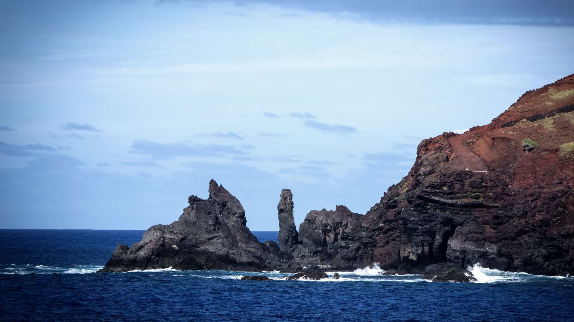 A scenic view of the rugged, volcanic coastline of Pitcairn Island in the South Pacific, with towering dark cliffs and unique rock formations jutting out of the deep blue water. 