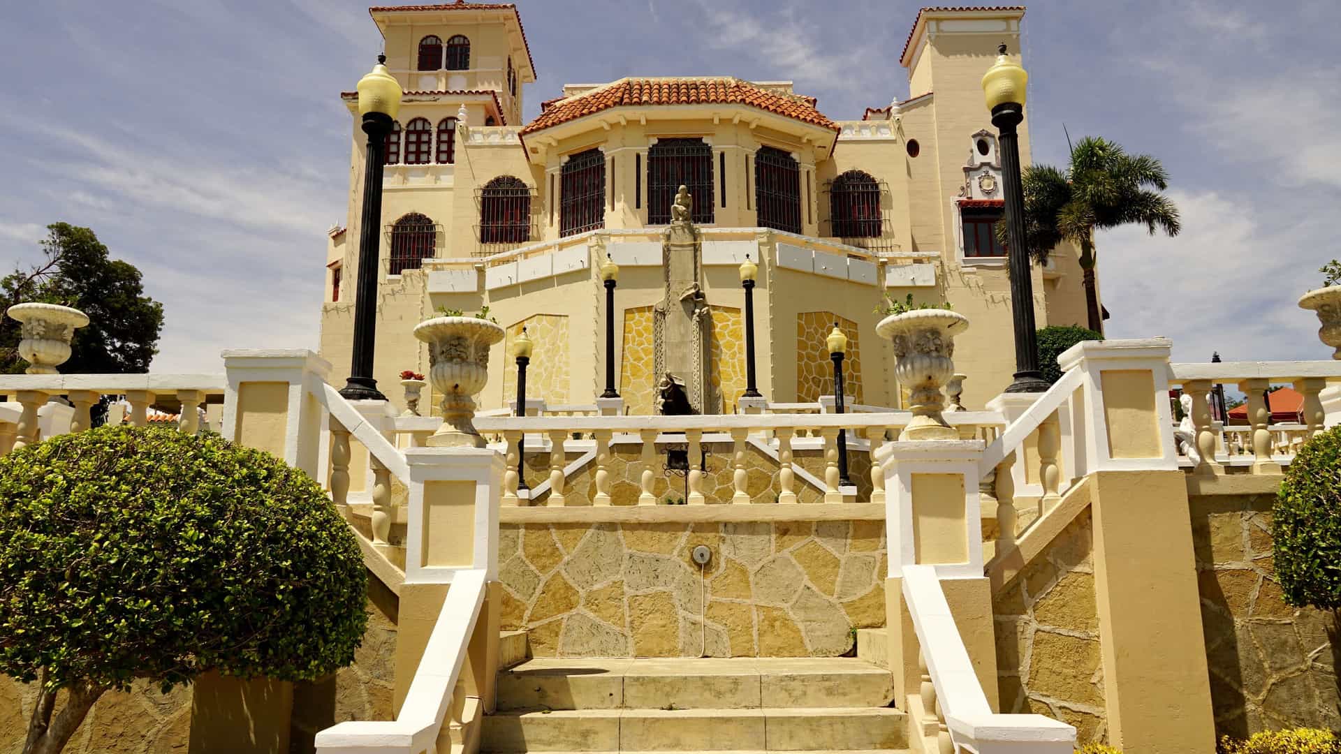 A view of the historic Serrallés Castle in Ponce, Puerto Rico, showing the grand staircase leading to the entrance of the Spanish Revival-style mansion with ornamental street lamps and a fountain.