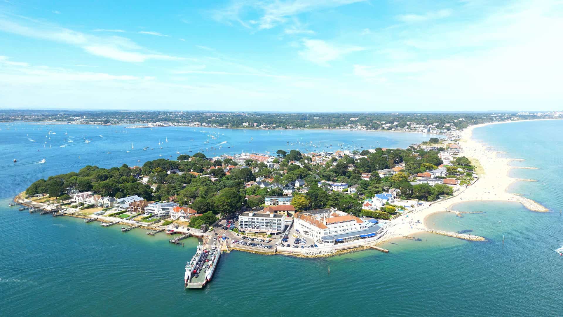 An aerial view of the scenic Sandbanks peninsula in Poole, Dorset, England, showing luxury homes, a ferry dock, and a sandy beach surrounded by the expansive blue waters of the harbor and the English Channel.