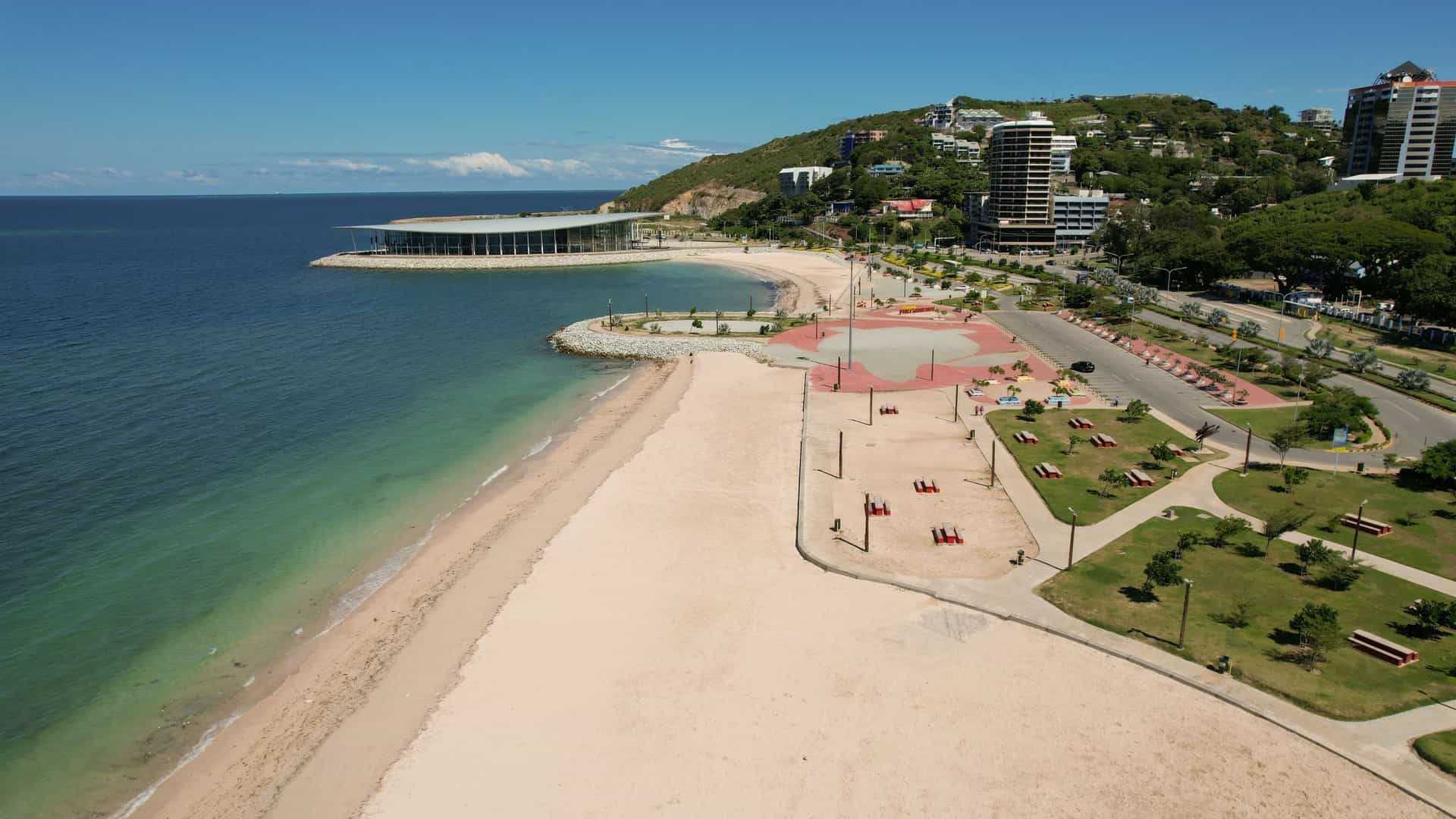An aerial view of the Port Moresby waterfront in Papua New Guinea, showing a large sandy beach, a newly developed park area with seating, and a modern building and high-rise apartments overlooking the harbor.