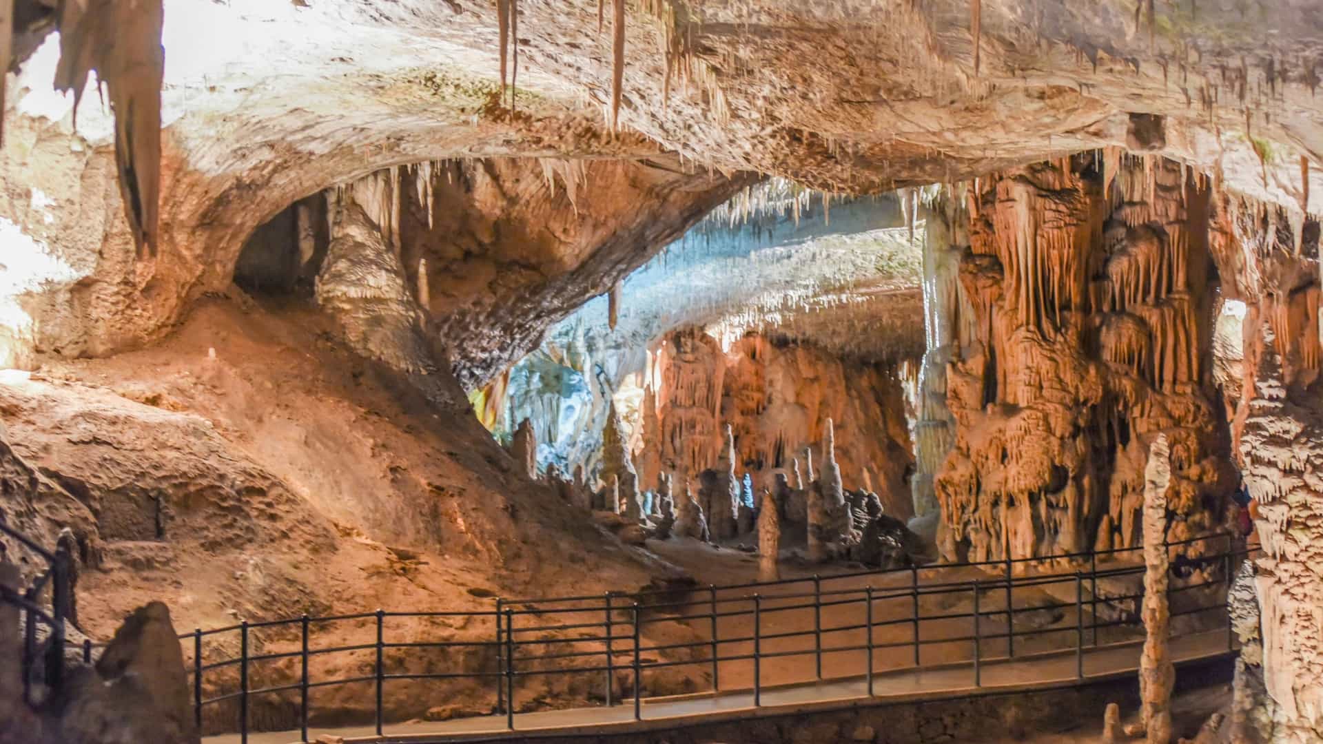 A view inside the illuminated Postojna Cave in Slovenia, with a paved walkway and stunning formations of stalactites, stalagmites, and rock columns.