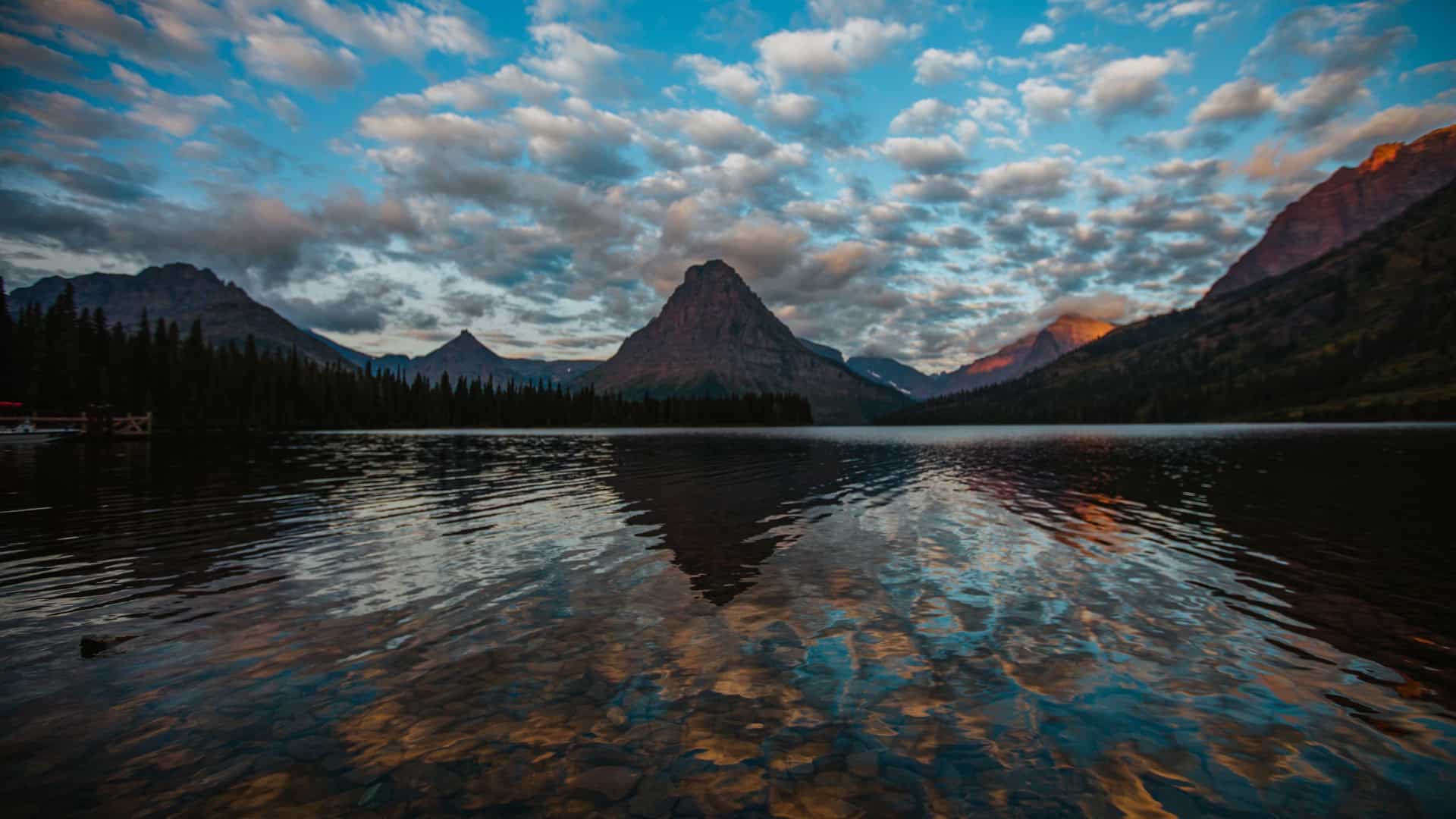 A dark, clear lake in Montana reflects a dramatic sunset sky and a large, symmetrical mountain peak surrounded by dense pine forests.