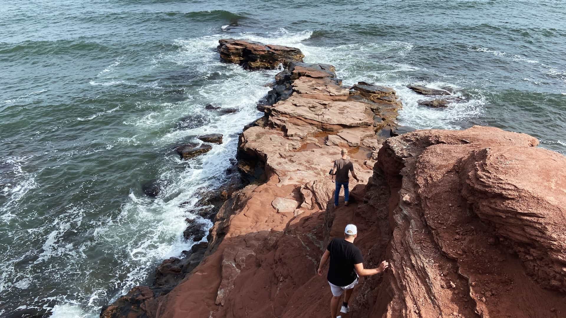 Two people walk down a rugged outcrop of dramatic, dark red sandstone cliffs that juts into the choppy, blue-gray ocean water.