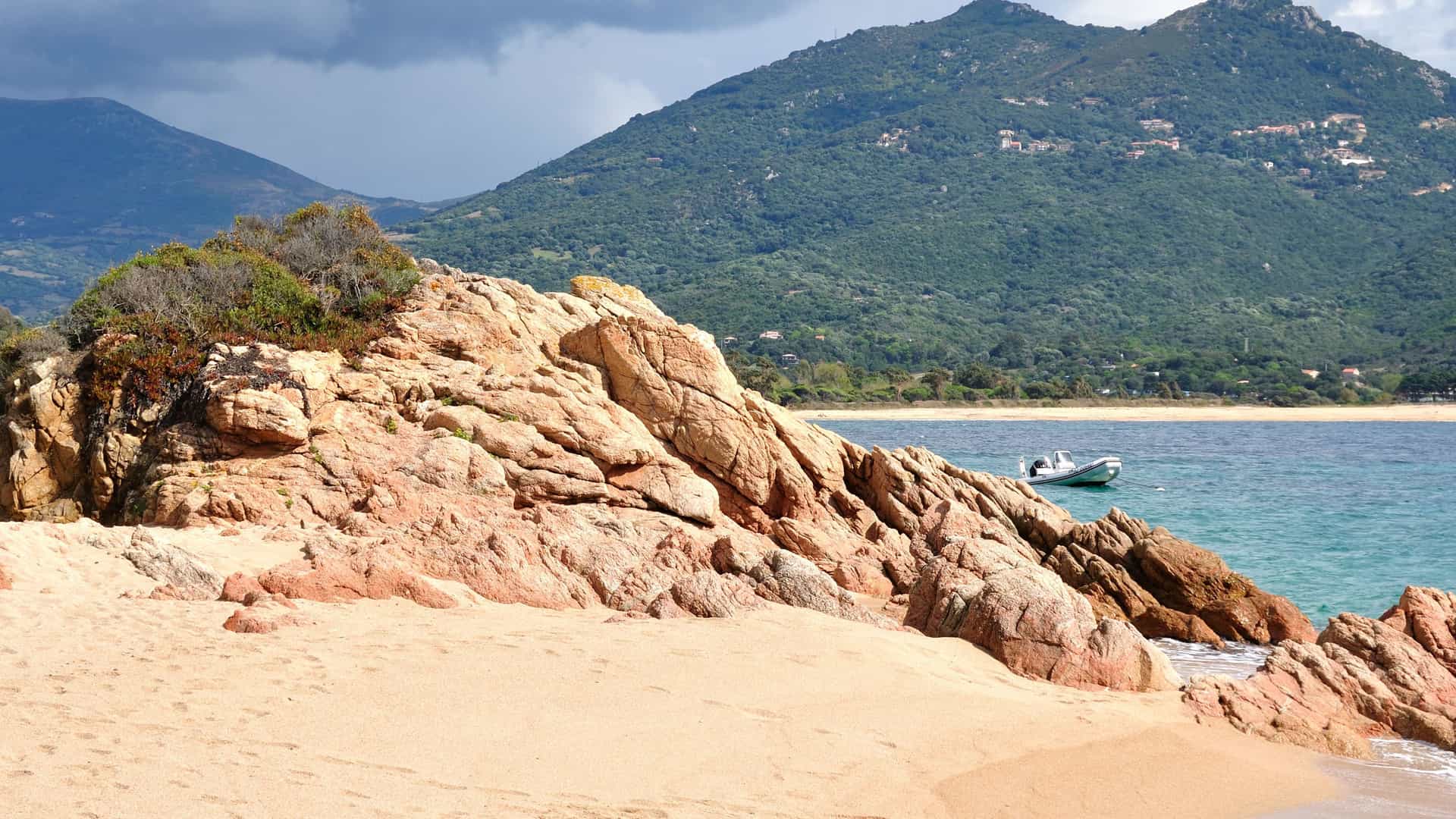 A serene beach scene in Propriano, Corsica, with a rocky coastline, a small boat on the turquoise water, and mountains covered with green trees in the background.