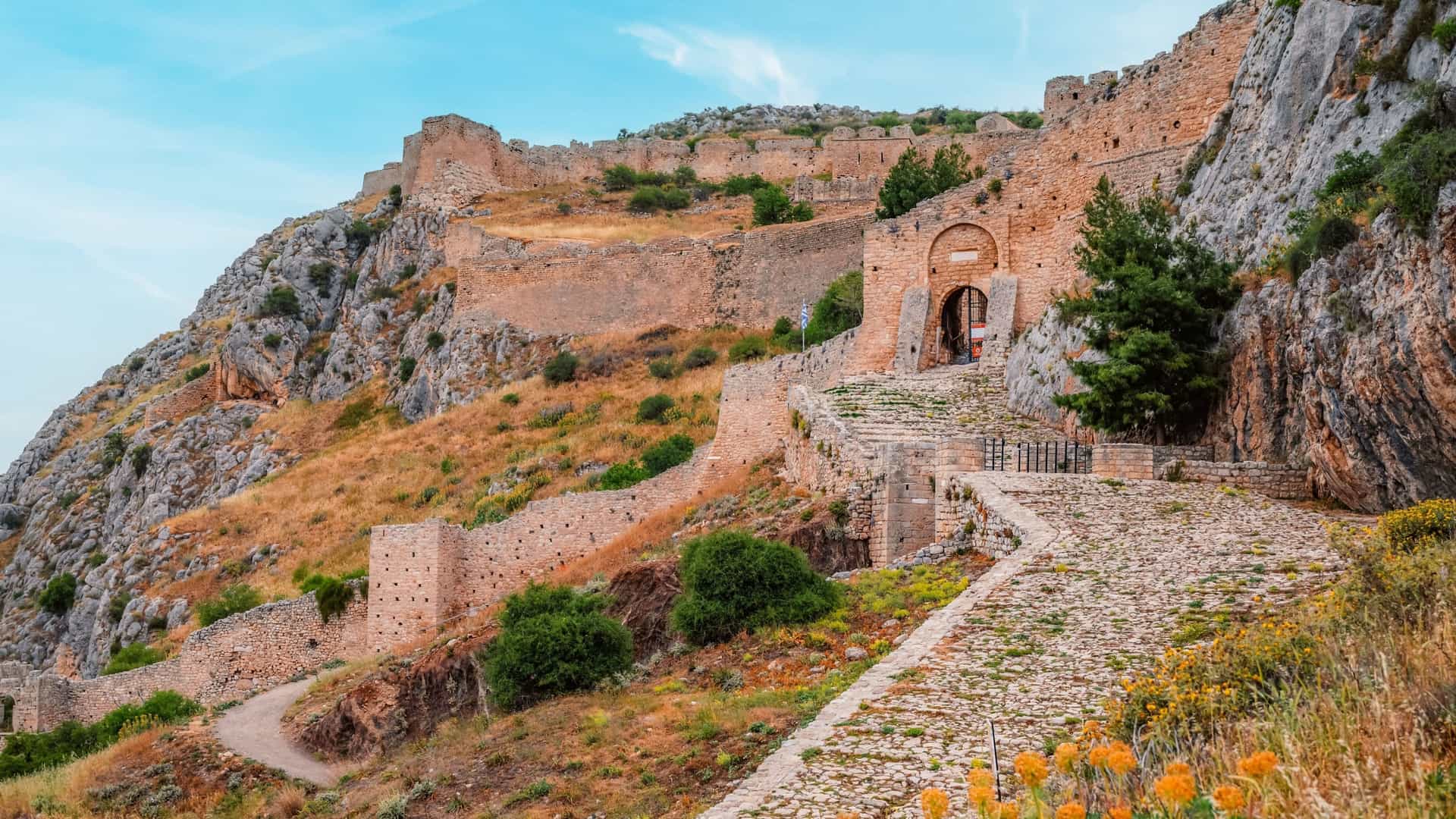 A panoramic shot of the historic Acrocorinth citadel in Greece, showcasing its ancient stone walls and a cobblestone pathway winding up a steep, rocky hill under a bright sky.