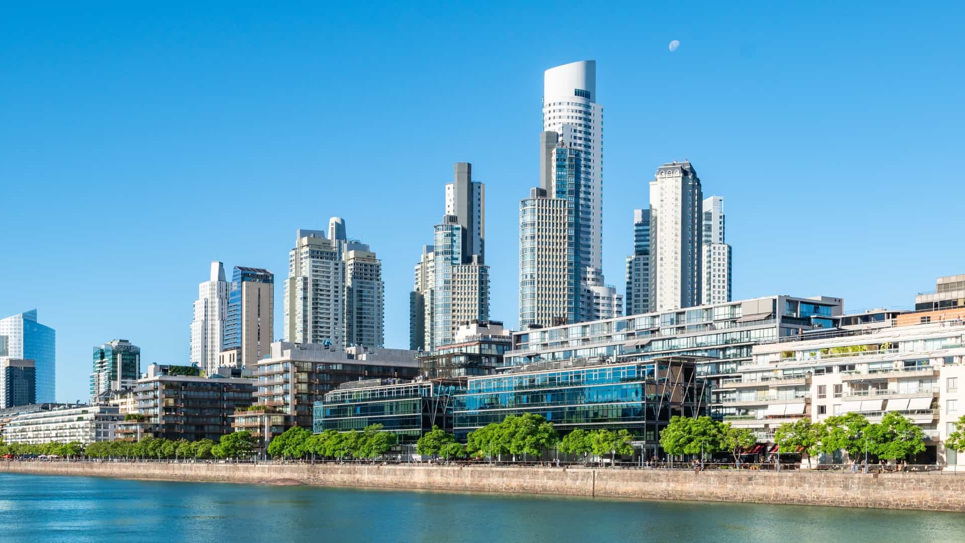 A row of modern, glass skyscrapers in the Puerto Madero district of Buenos Aires line a waterfront on a clear day.
