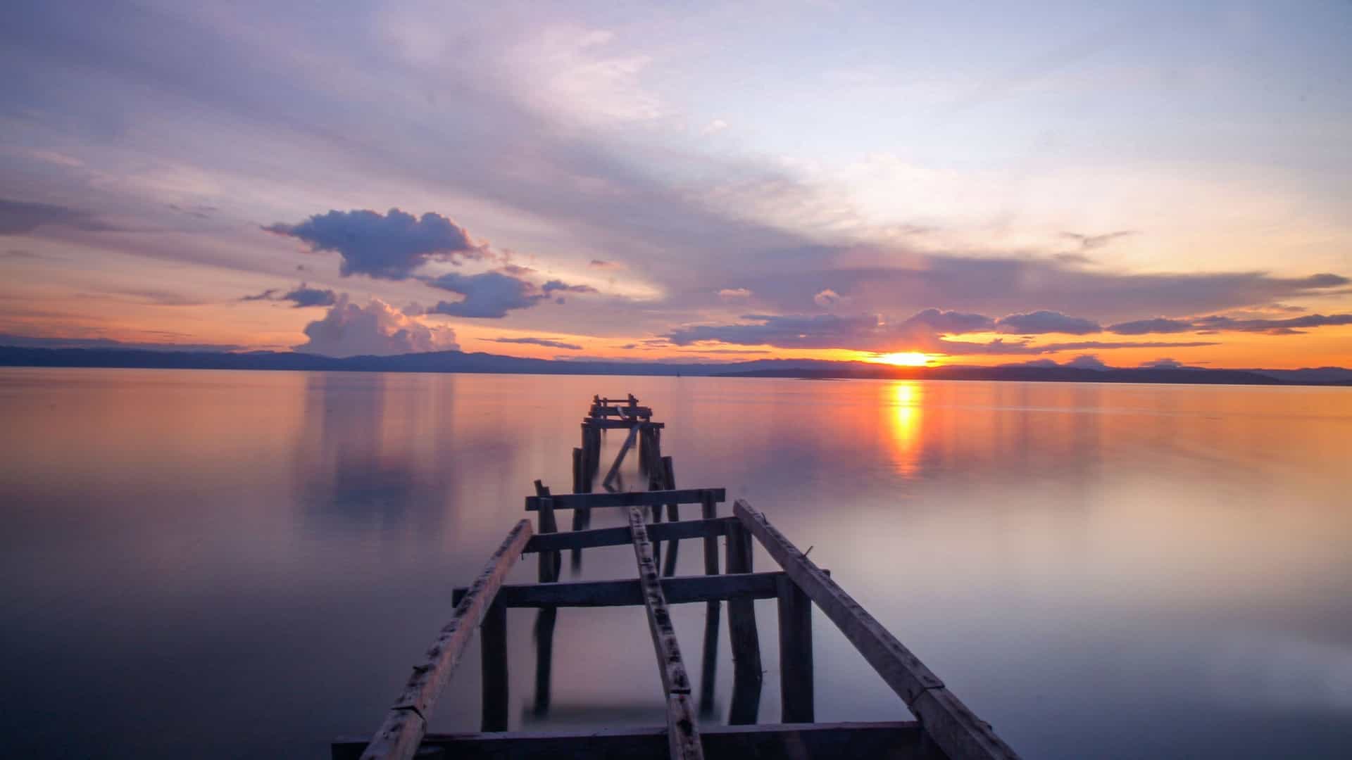 A beautiful sunset shot over the still, reflective water, with the sun setting behind distant mountains and the remains of an old, rustic wooden pier in the foreground at Pulau Rouw in Raja Ampat, Indonesia.