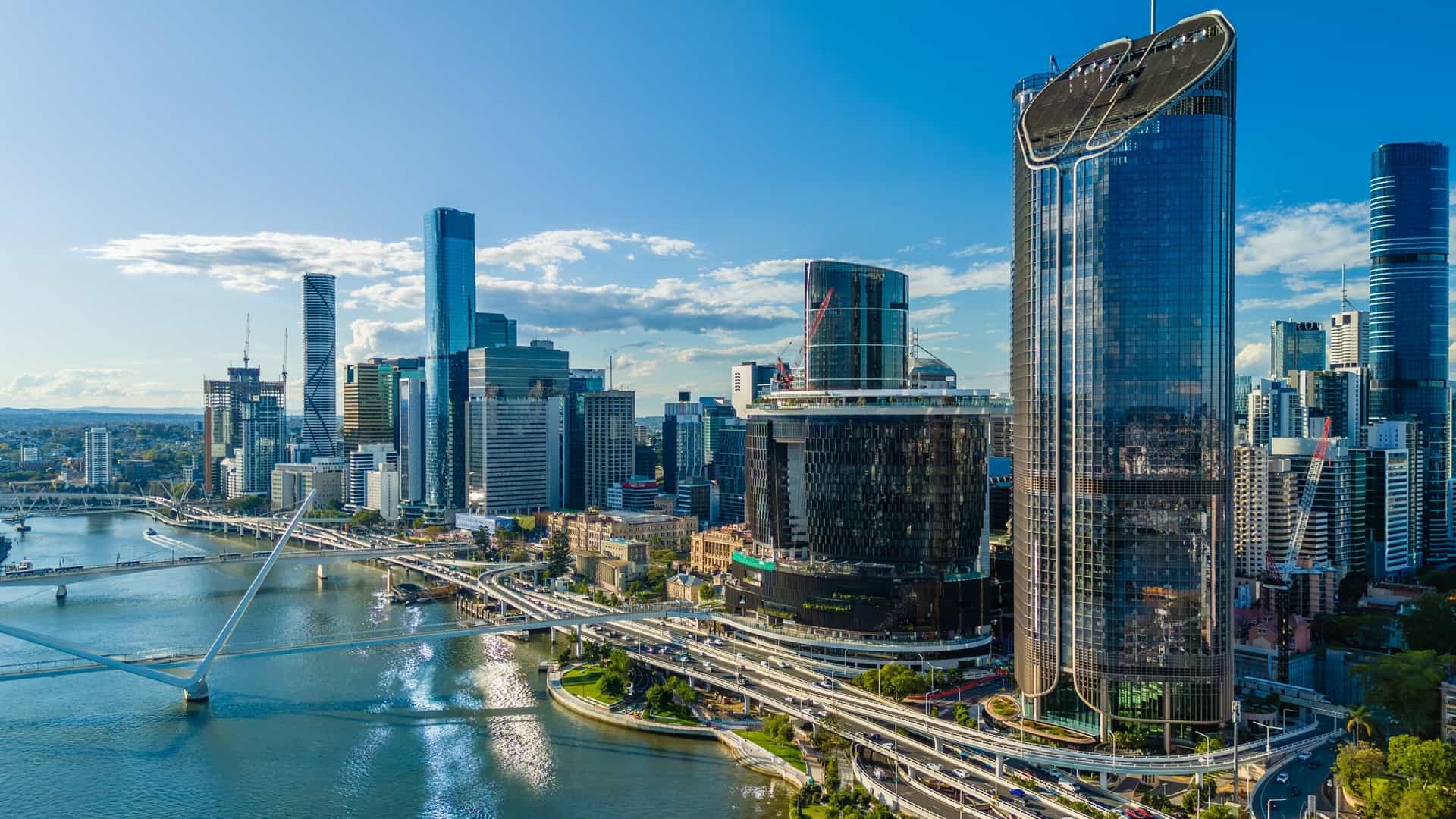 An aerial view of the Brisbane skyline features a river, a bridge, and numerous towering, modern, glass skyscrapers under a blue sky.