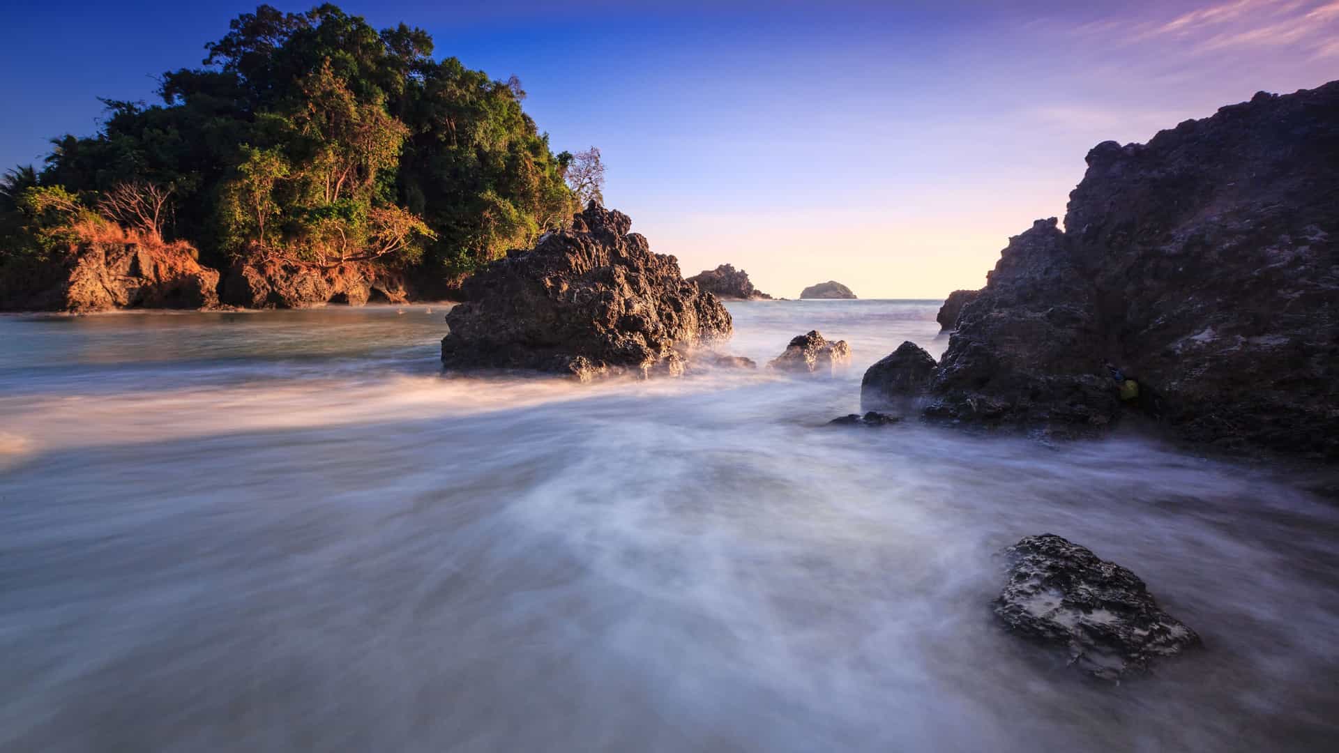 A dreamy long-exposure shot of the coastline in Manuel Antonio National Park near Quepos, Costa Rica, with a tranquil, misty ocean flowing between large, jagged rocks and a lush green headland in the distance at sunset.