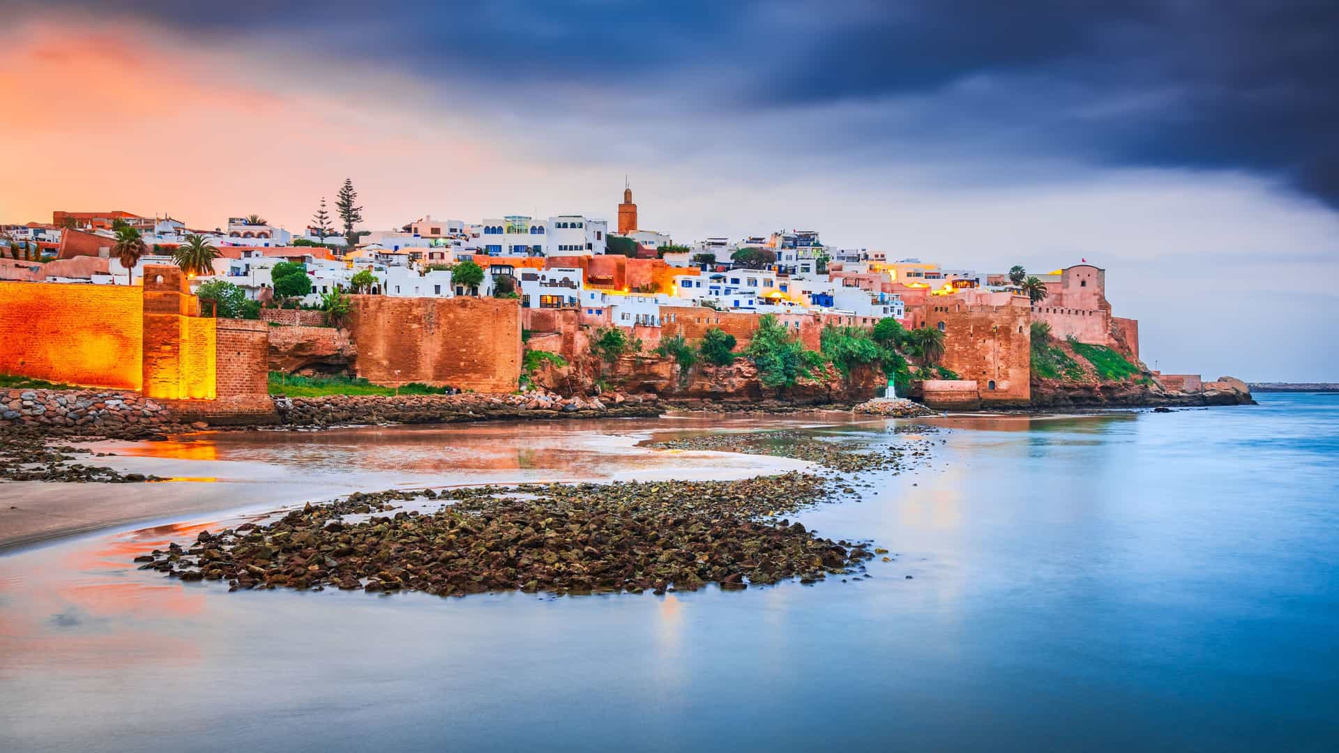 "A scenic view of the Kasbah of the Udayas in Rabat, Morocco, at dusk. The ancient, fortified walls and white-and-blue buildings are warmly lit, overlooking a calm body of water that reflects the vibrant orange, purple, and blue hues of the sunset sky.  "