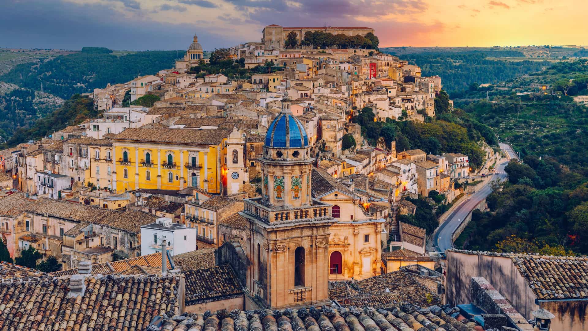 "An elevated view of the historic Baroque town of Ragusa Ibla in Sicily, Italy, showing a dense cluster of buildings and terracotta rooftops on a steep hillside. In the foreground, a prominent church with a striking blue dome and clock tower stands out, with the warm colors of a sunset filling the sky above.  "