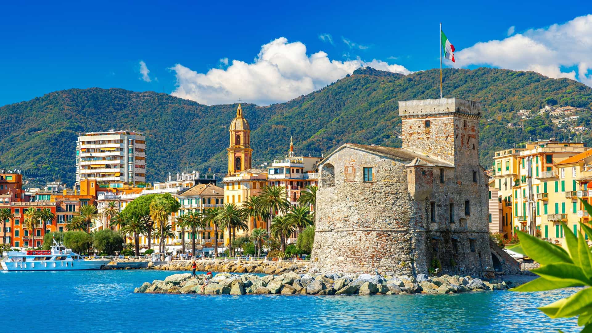 A small, centuries-old stone fort stands on the waterfront of Rapallo, with colorful buildings and lush green mountains in the background.