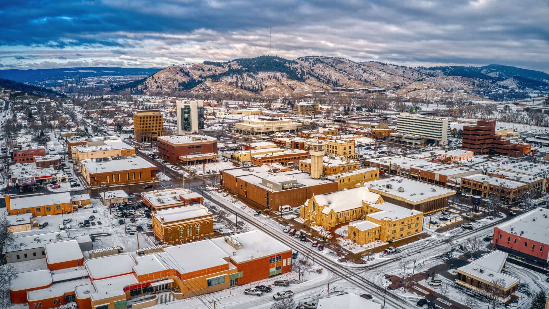 "An aerial view of downtown Rapid City, South Dakota, covered in a light blanket of snow during winter. The buildings, a mix of modern and older low-rises, have rooftops dusted with white, and the streets are visible as dark lines. The city is nestled against the backdrop of snow-covered Black Hills, with evergreen trees and patches of white snow on the slopes under a dramatic, cloudy sky.  "