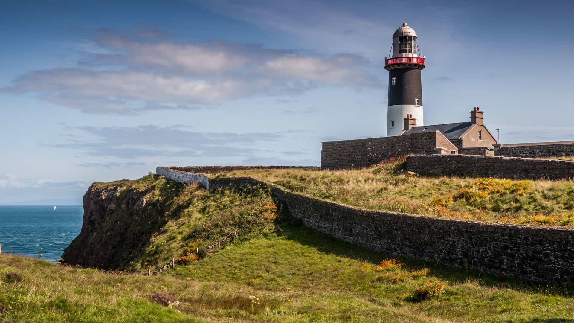 "A scenic view of the Rathlin West Lighthouse on Rathlin Island, Northern Ireland. The black and white lighthouse, with its red top, is situated on a lush green cliff, with low, winding stone walls defining the grounds. The lighthouse overlooks the deep blue sea under a partly cloudy sky.  "