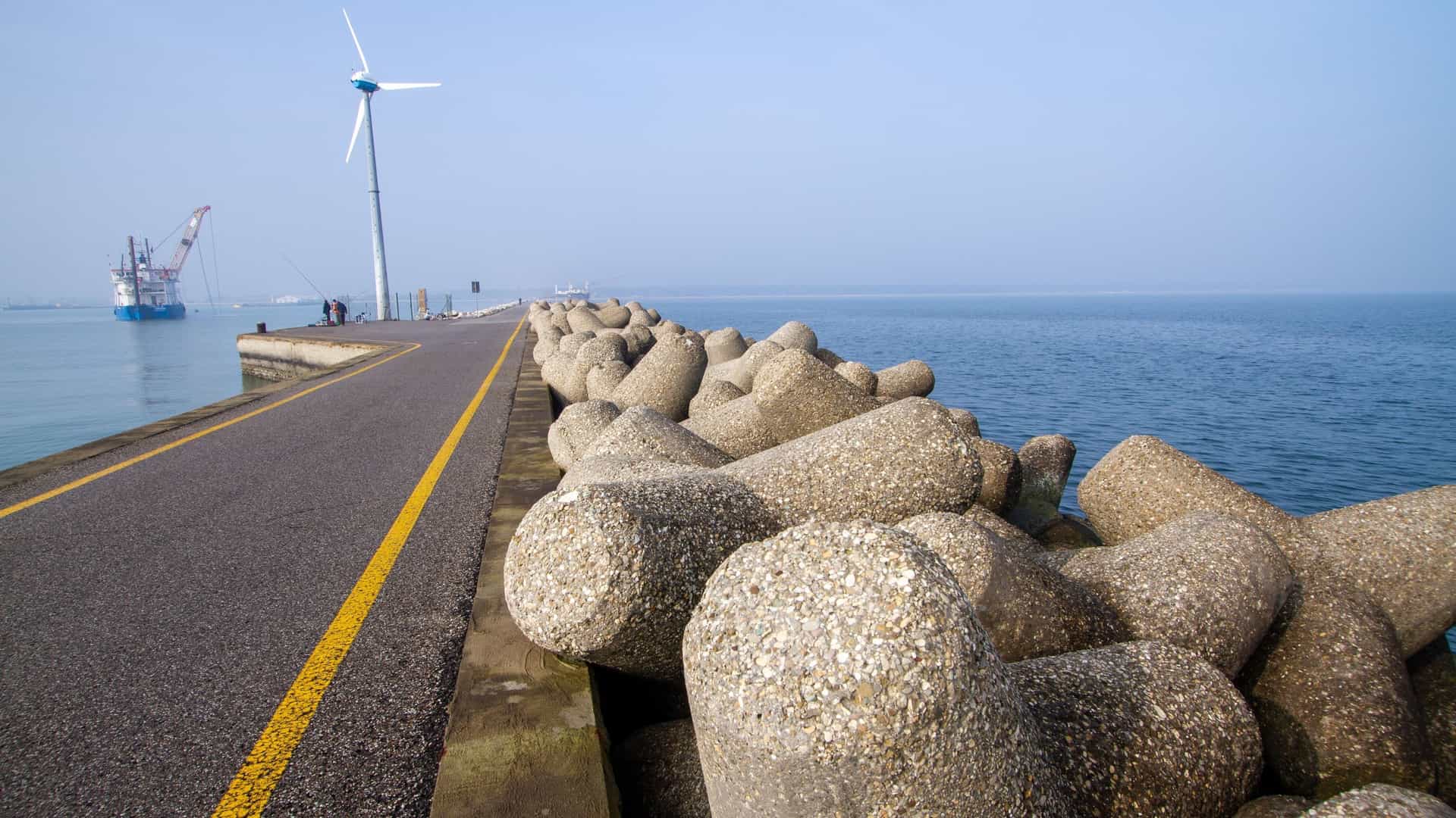 A long concrete pier or jetty stretches out from the coast into the calm blue waters of the Adriatic Sea at Porto Corsini, which is located within the Regional Park of the Po Delta in Ravenna, Italy. The sky is bright with fair weather.