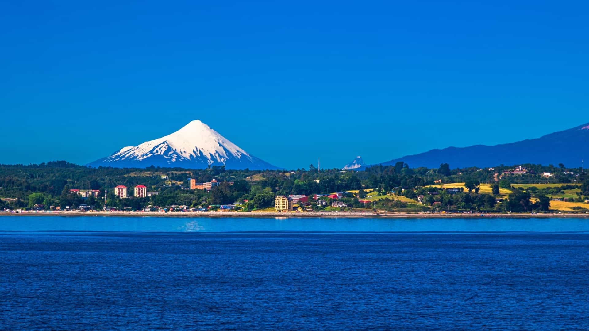 A beautiful landscape shot of the Reloncavi Sound in Chile, with a snow-capped Osorno volcano and lush green shoreline visible across the tranquil blue water.