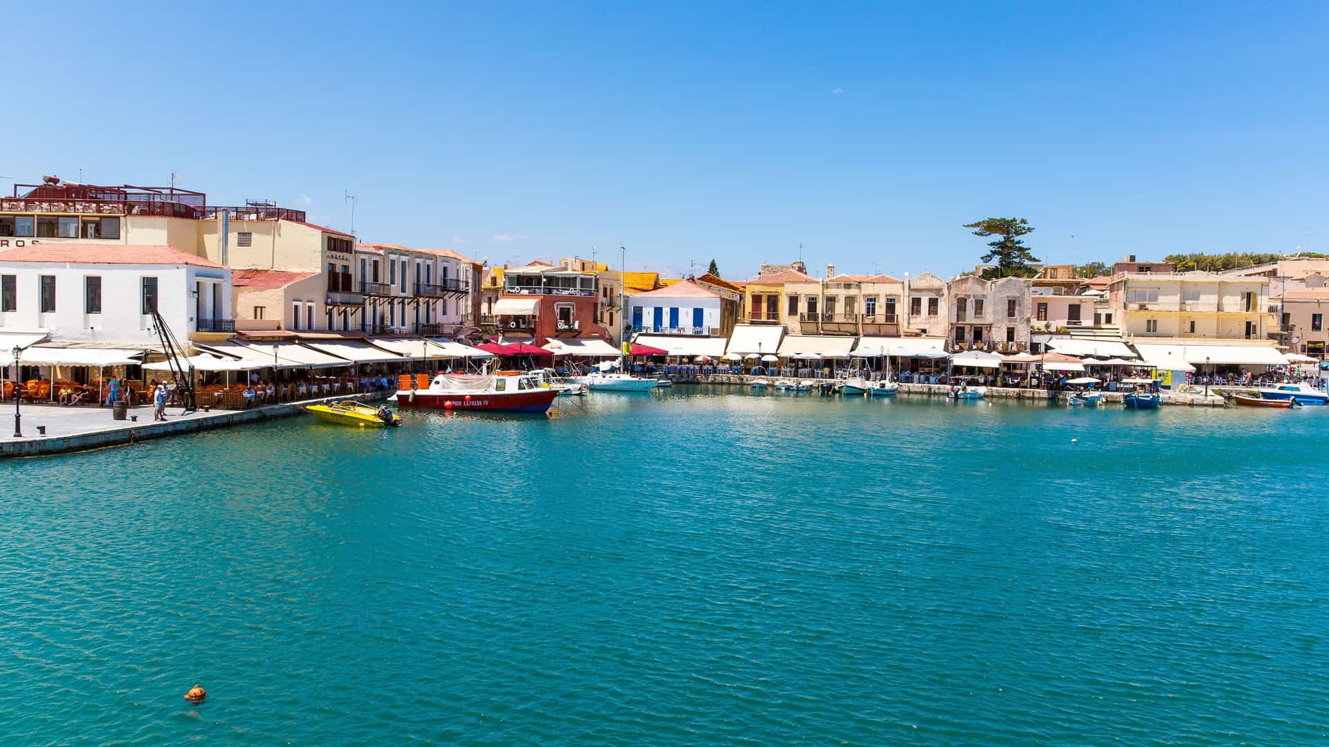 A colorful photo of the Rethymnon harbor in Crete, Greece, with boats and waterfront buildings with awnings, all under a bright blue sky.