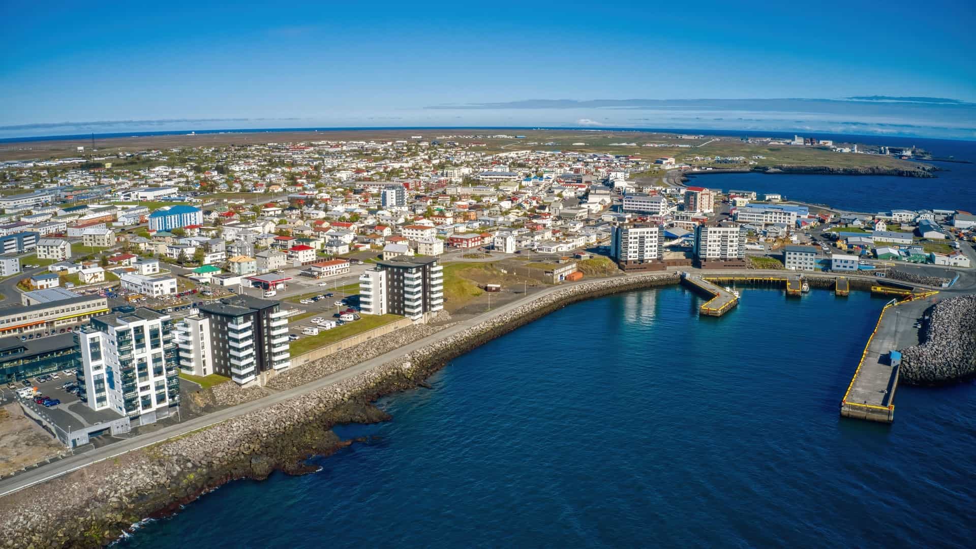 An aerial view of Reykjanesbær, Iceland, showing the coastal town and its harbor with modern and traditional buildings, set against a backdrop of a clear blue sky and the Atlantic Ocean.