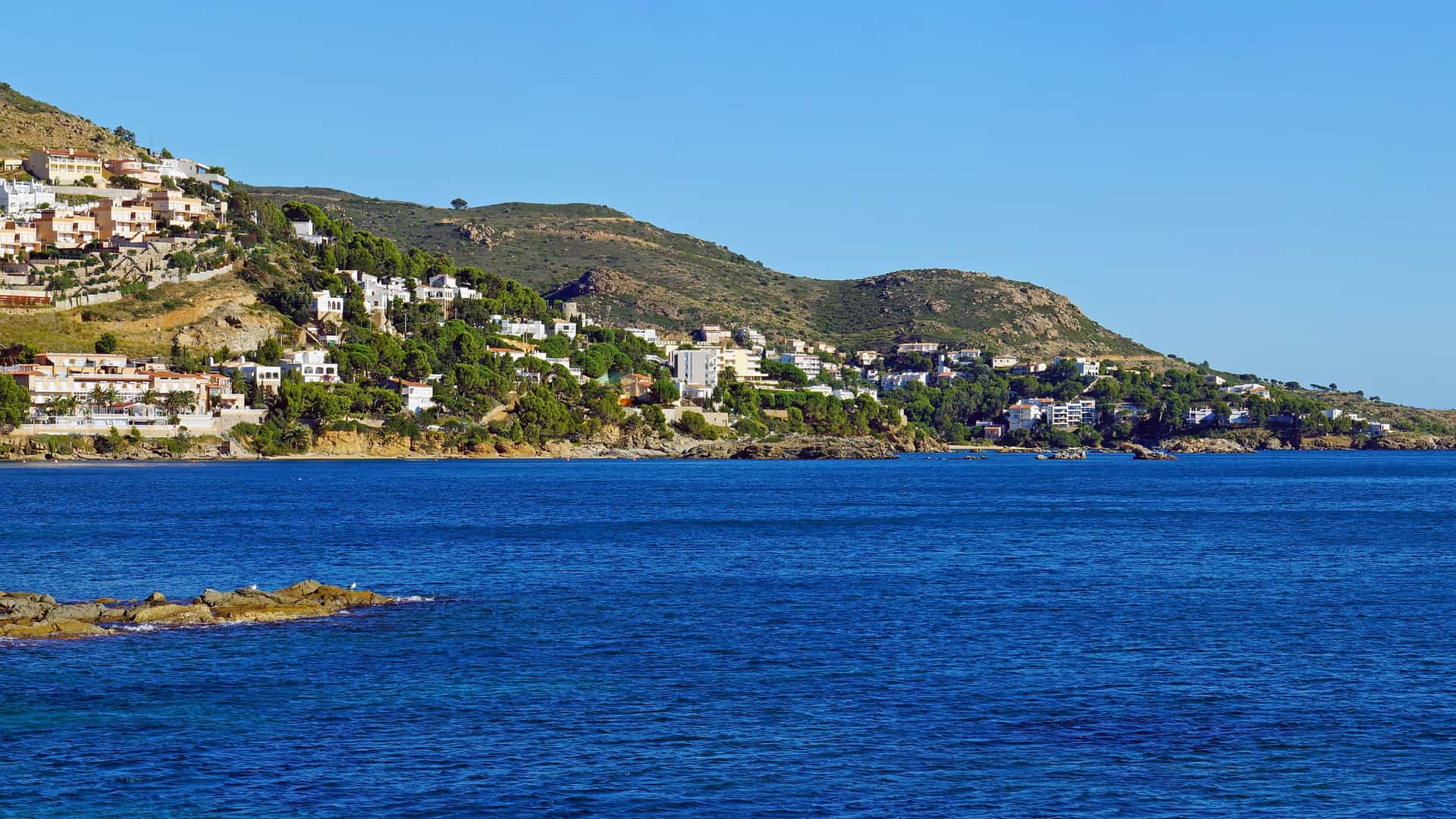 A wide shot of Roses, Spain, showing the Mediterranean coastline with a small town of white buildings climbing up a green hillside overlooking the deep blue sea.