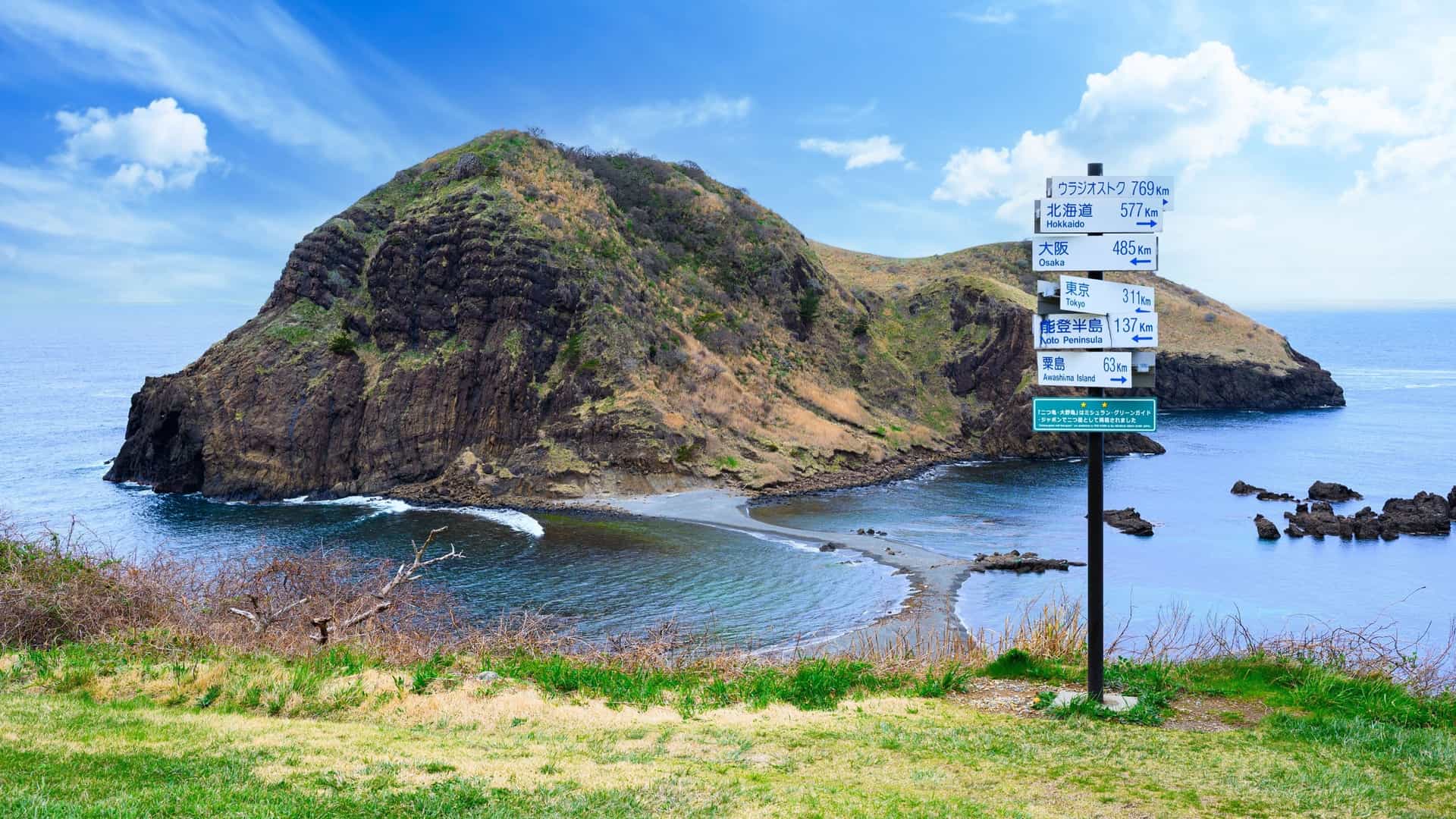 A destination signpost stands before a volcanic hill rising from the bay on the scenic coastline of Sado Island, Japan.