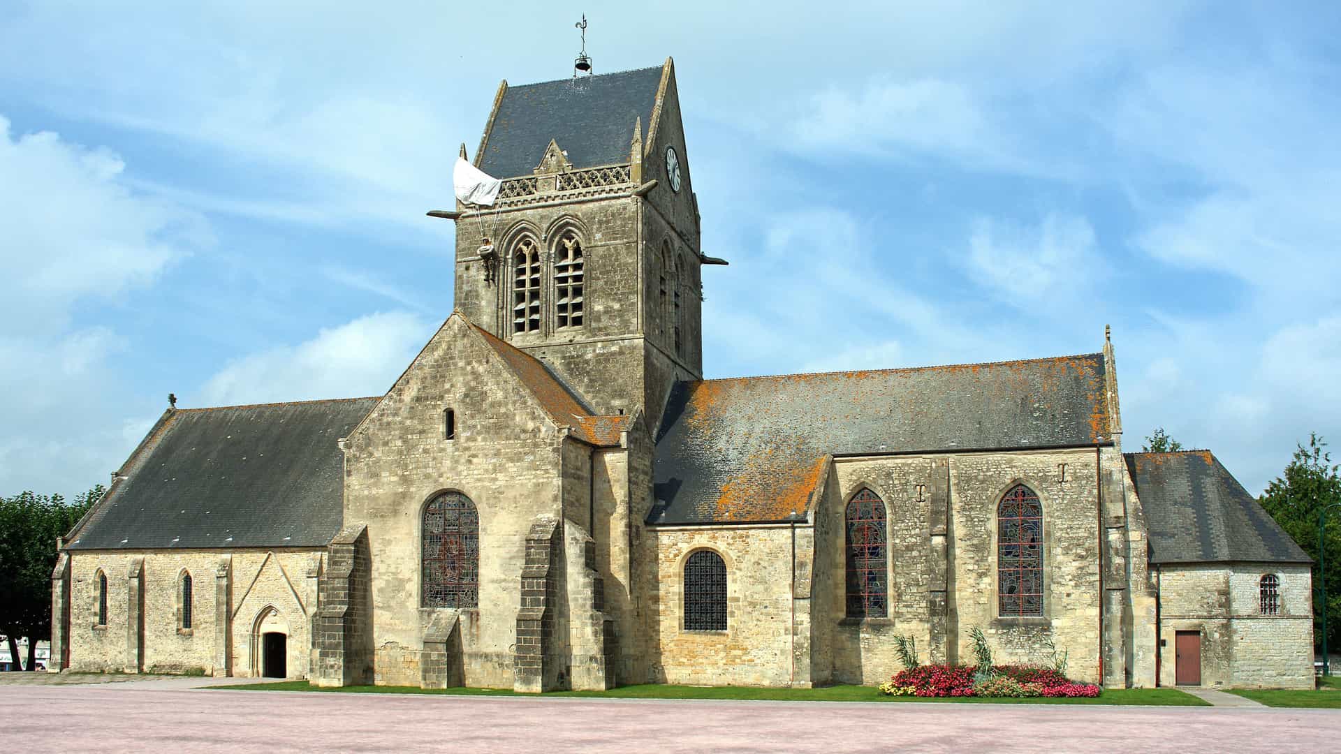 A wide-angle view of the historic Sainte-Mère-Église church in Normandy, France, famous for its role in the D-Day landings, with a parachute and figure hanging from the steeple.