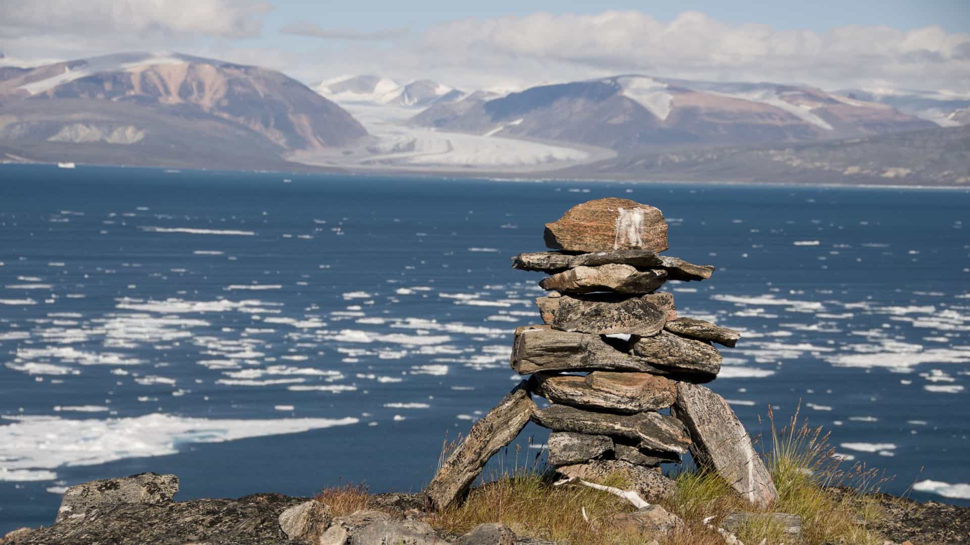 A striking rock cairn, or inukshuk, in the foreground overlooking the cold, ice-filled waters of Sam Ford Fjord, with snow-capped mountains and glaciers in the background.