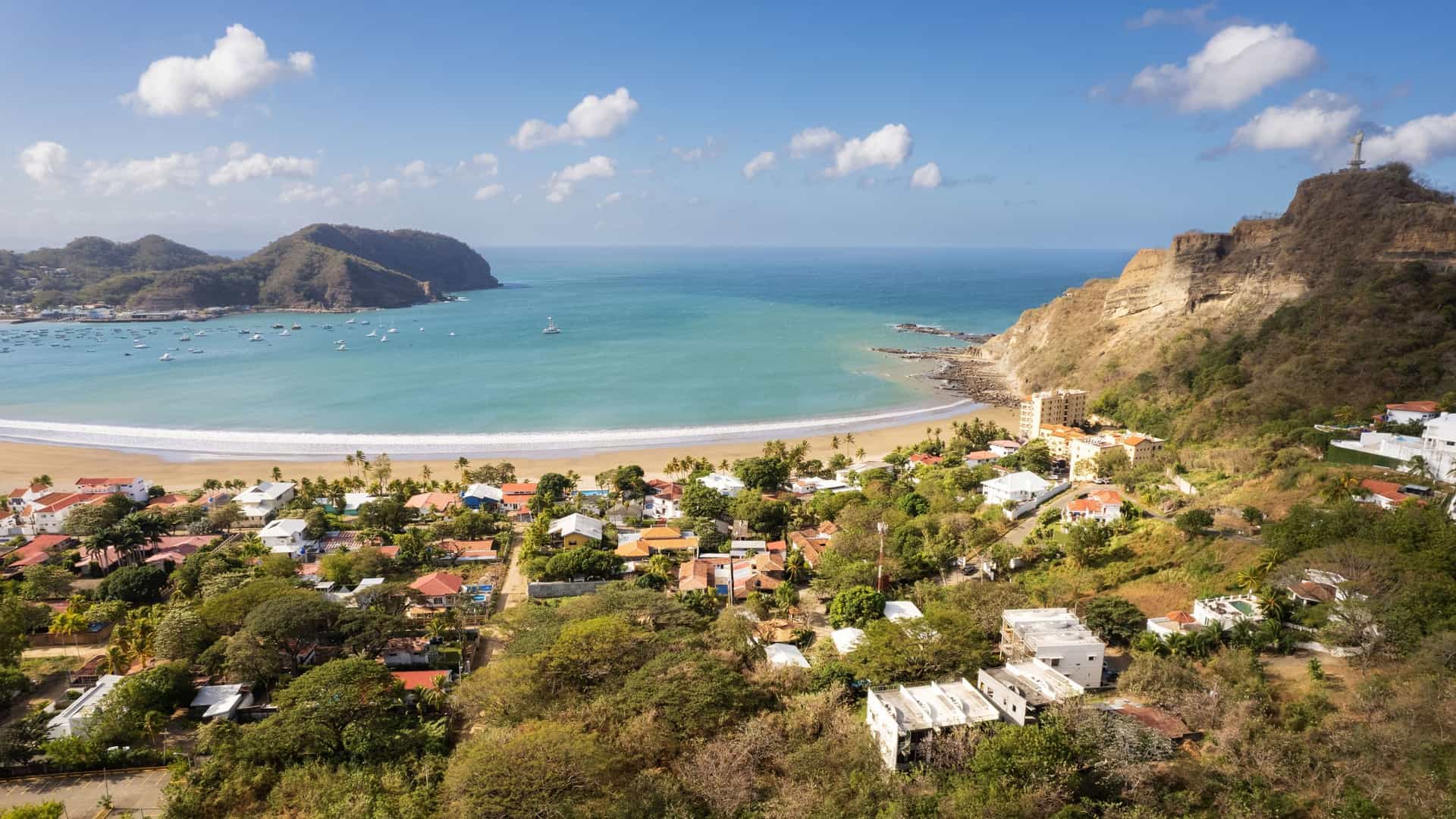 A beautiful aerial view of the horseshoe-shaped bay of San Juan del Sur, Nicaragua, with a sandy beach, a coastal town, and a statue of Christ the Redeemer on the hill under a bright blue sky.