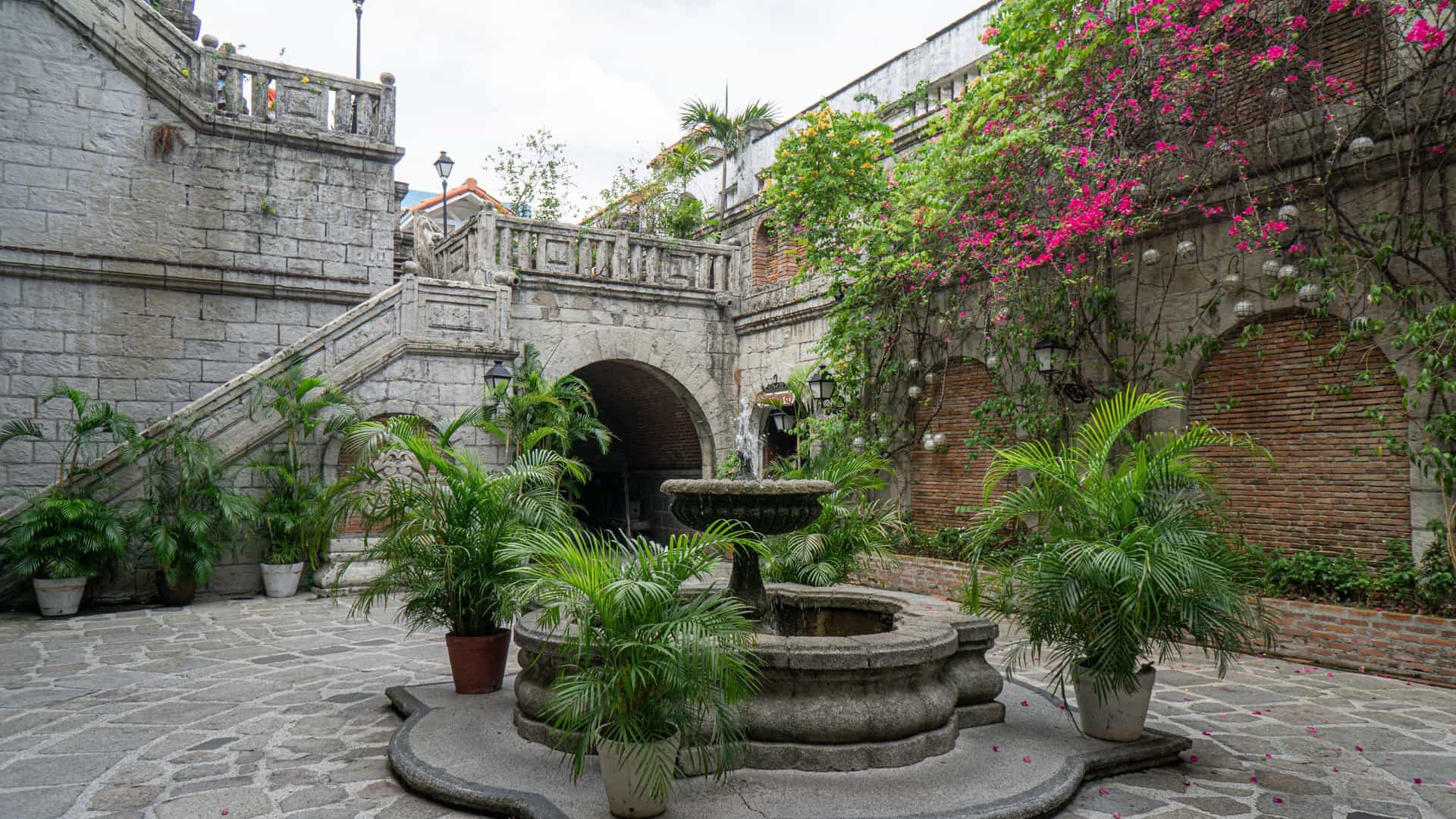 A stone courtyard with a central fountain, bordered by ivy-covered brick walls and staircases in San Luis, The Philippines.