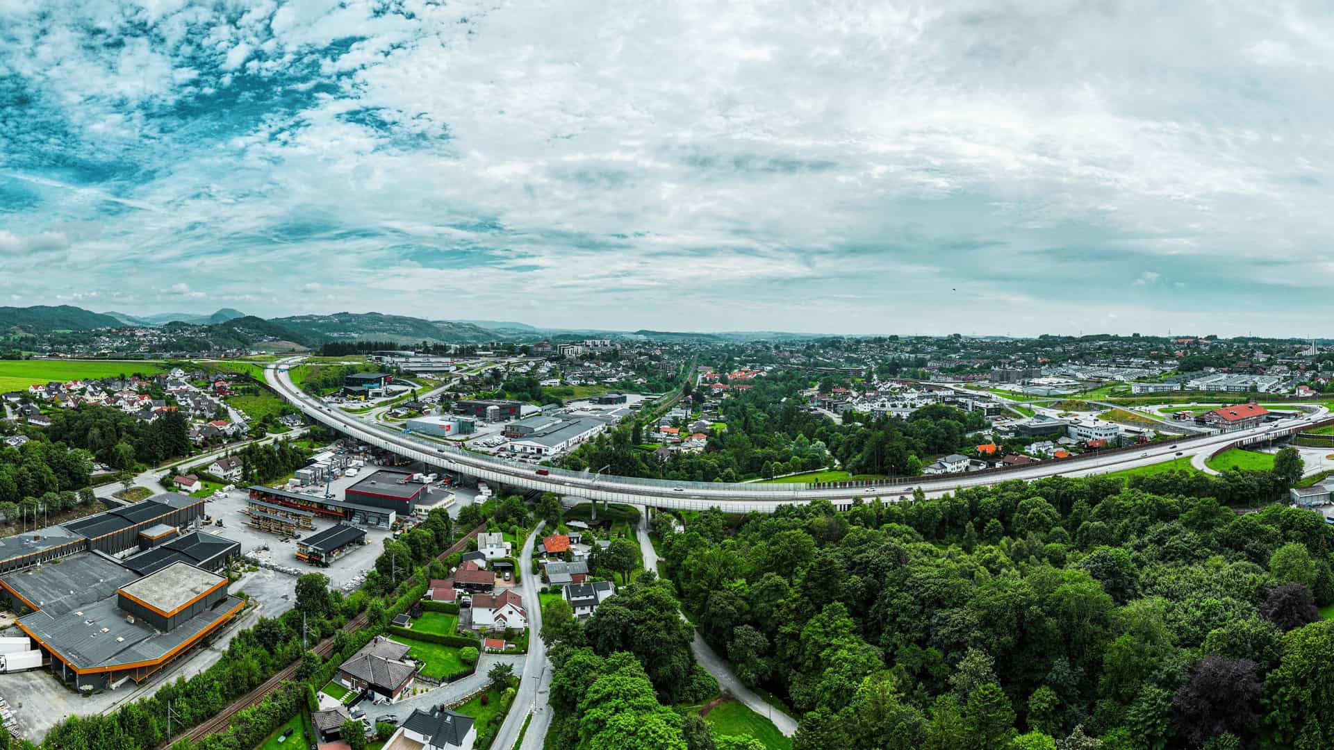 A panoramic view of the cityscape of Sandnes, Norway, situated at the end of the Gandsfjord. The city features a blend of modern and traditional Scandinavian architecture concentrated along the waterfront, with residential areas extending up the surrounding lush green hills and mountains. The wide shot highlights the contrast between the urban development and the natural fjord landscape.
